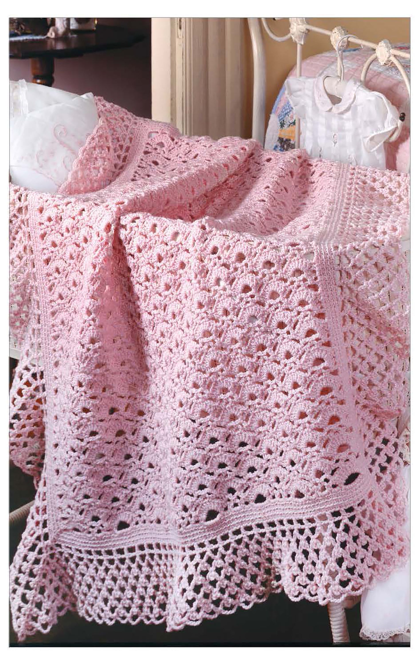 A light pink, intricately crocheted baby blanket with scalloped edges and delicate patterns, inspired by classic designs from Leisure Arts’ The Little Encyclopedia of Crocheted Baby Blankets, draped over a white chair.