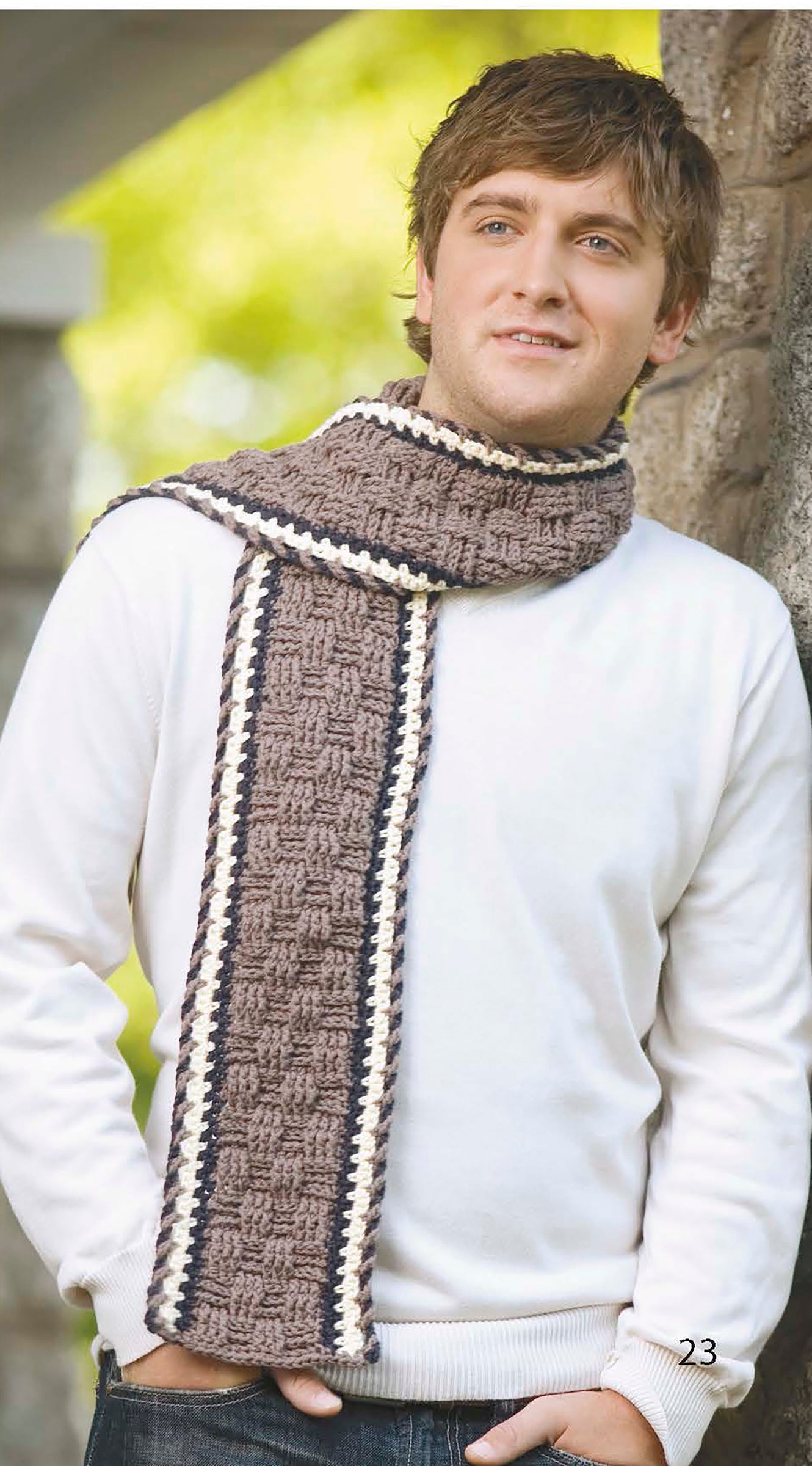 A man smiles outdoors, wearing a white sweater and a knitted scarf in brown, white, and dark gray, featuring a basketweave pattern inspired by beginner designs from Leisure Arts’ "The Little Encyclopedia of Crocheted Scarves" digital download.