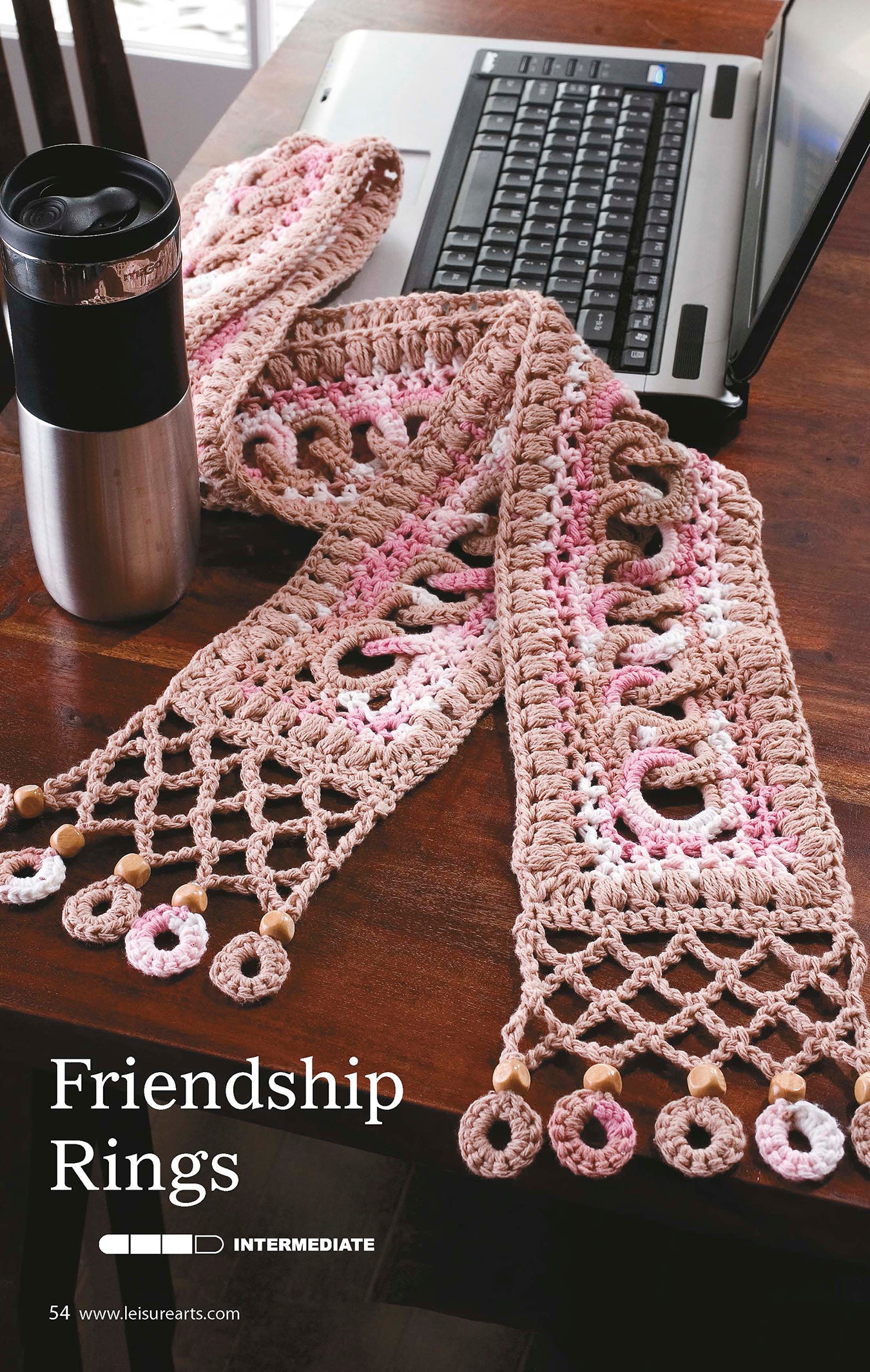A pink and beige crocheted scarf with circular patterns is draped on a wooden table beside a laptop and travel mug. Text reads The Little Encyclopedia of Crocheted Scarves by Leisure Arts, featuring unique intermediate scarf designs.