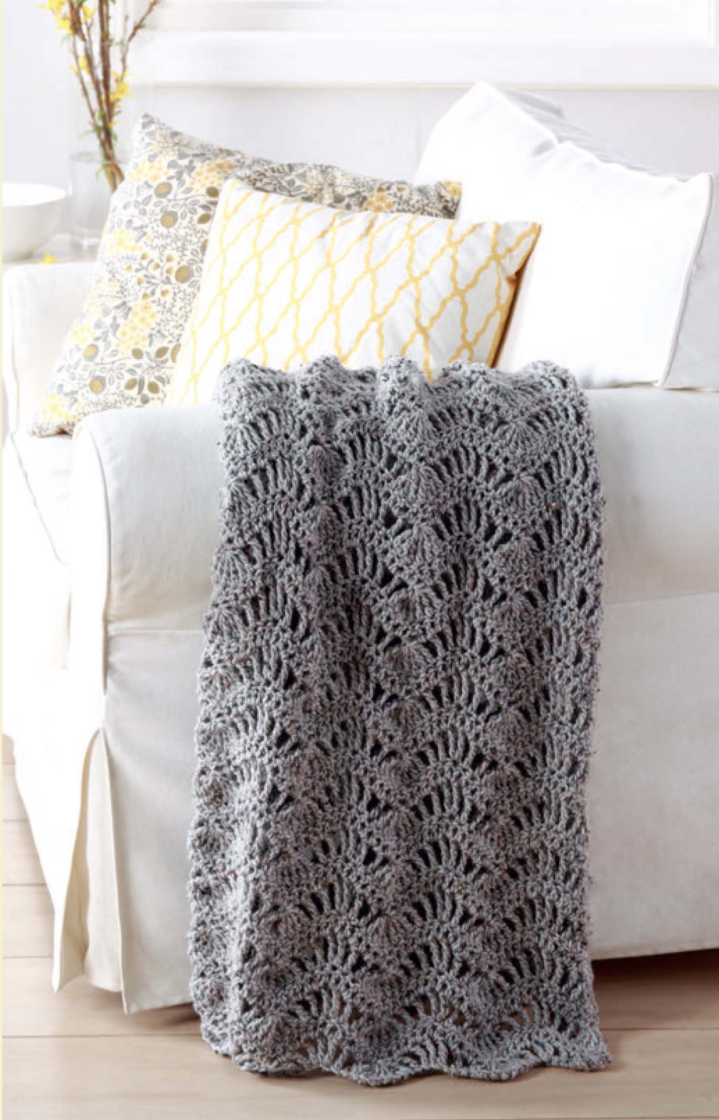 A cozy gray crocheted afghan, inspired by Leisure Arts' "Make In A Weekend—Afghans To Crochet" digital download, is draped over a white sofa with patterned pillows. In the background, a vase of branches sits on the side table.