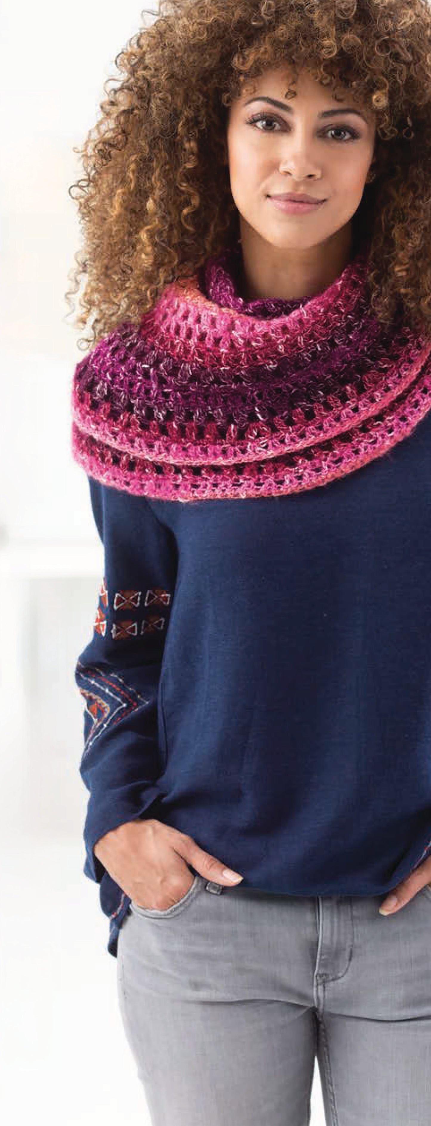 A woman with curly hair smiles slightly at the camera indoors, wearing light gray jeans, a blue sweater with patterned sleeves, and a chunky pink and purple cowl made from "Shawls & Vests—Crochet 7 Bohemian Designs" by Leisure Arts.