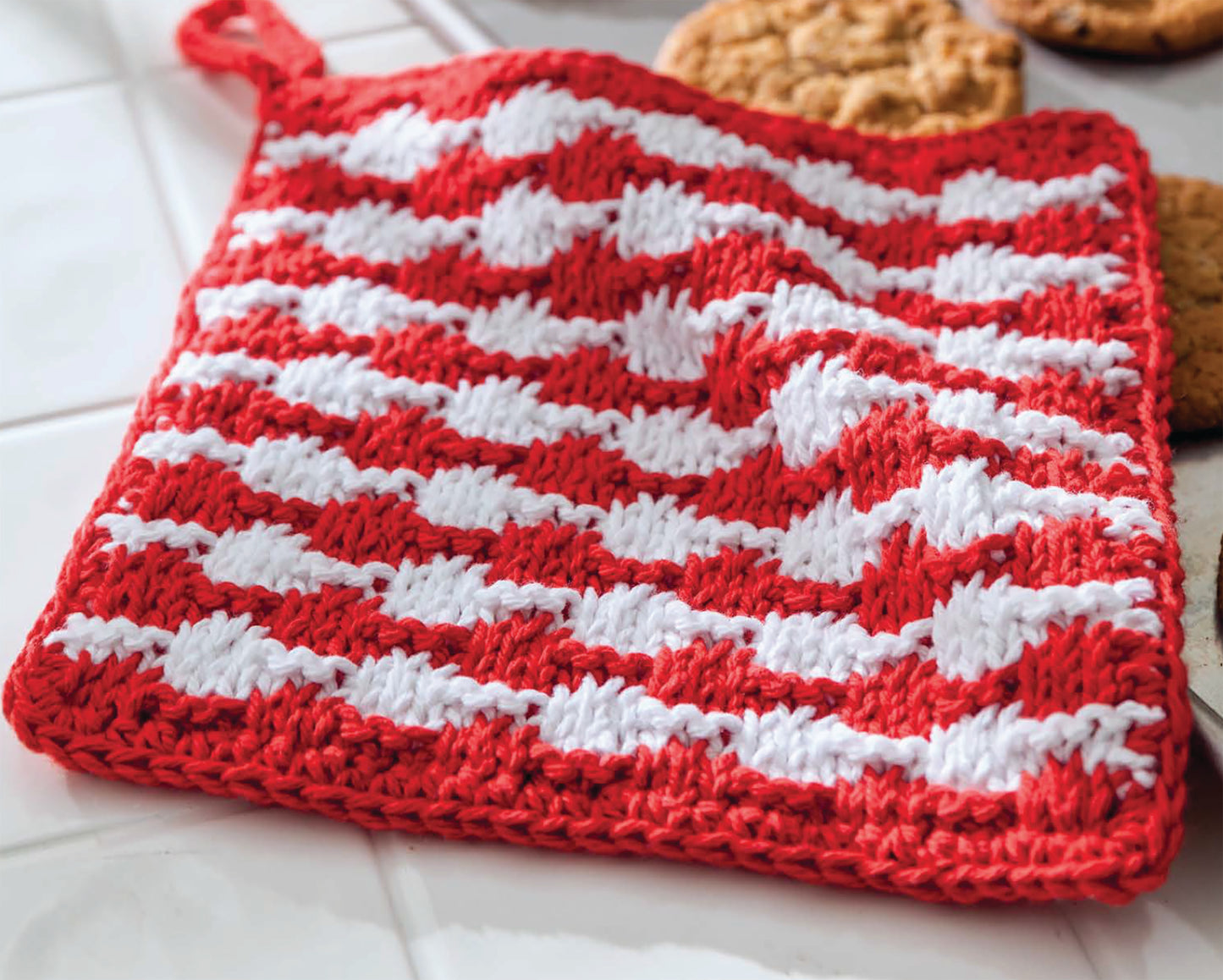 A red and white crocheted pot holder from Leisure Arts’ Make in a Weekend Potholders and Dishcloths lies on white tiles near cookies, featuring a striped, textured pattern perfect for handmade kitchen enthusiasts.