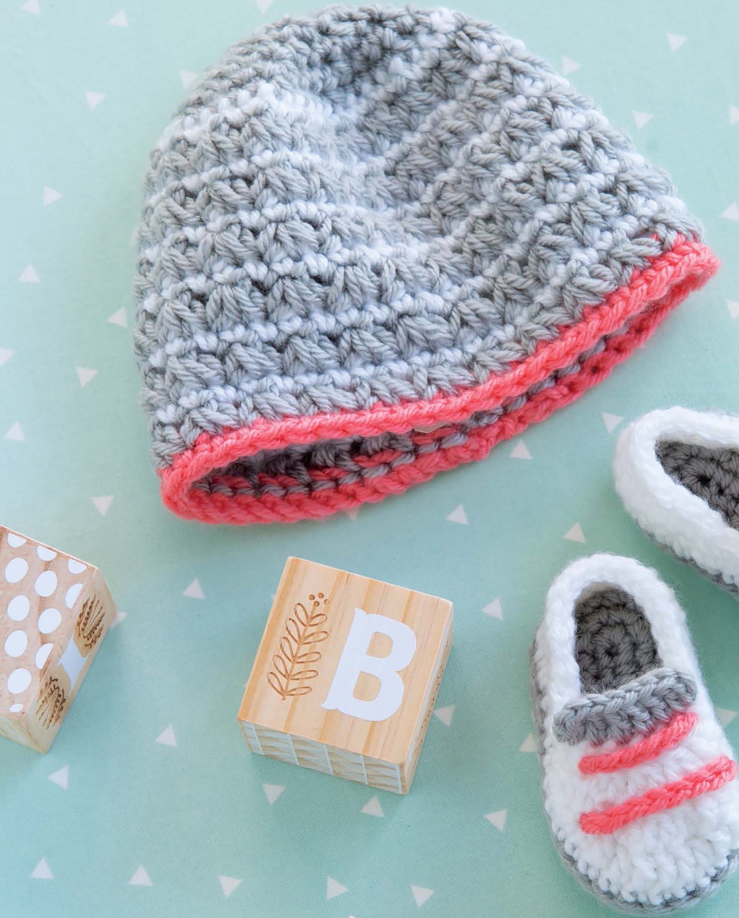 Leisure Arts Infant Boots & Hats set, featuring a gray crocheted baby hat with pink trim and matching booties, is displayed with wooden alphabet blocks on a mint green background with white triangles.