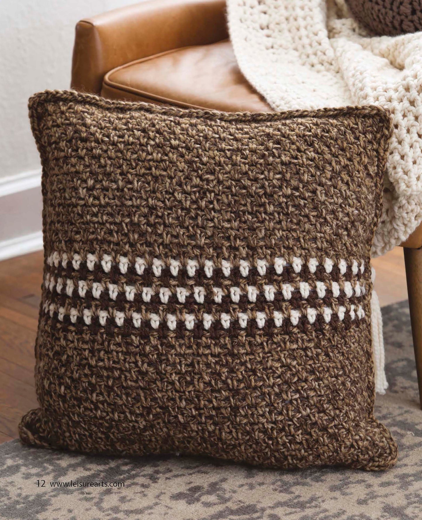 A brown crocheted pillow with textured patterns and a cream stripe sits on a tan leather chair with a chunky knit blanket, beautifully showcasing an easy project from Quick Crochet Home Decor by Leisure Arts.