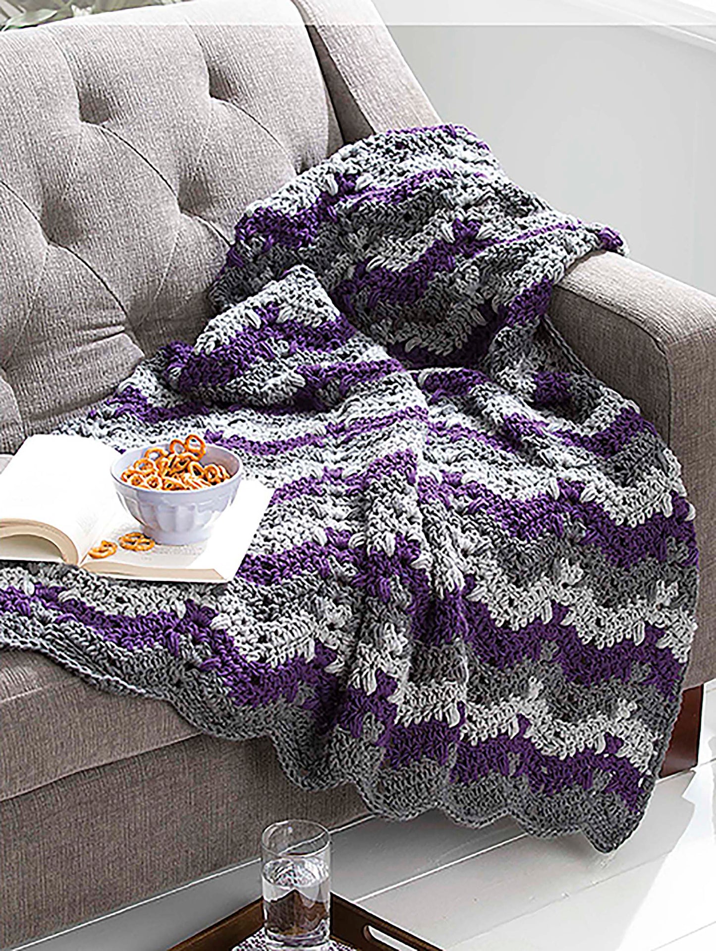 The Leisure Arts "Make in a Weekend Textured Lap Throws" digital download features a cozy gray, purple, and white striped crochet blanket draped over a beige sofa beside an open book, pretzels, and a glass of water.
