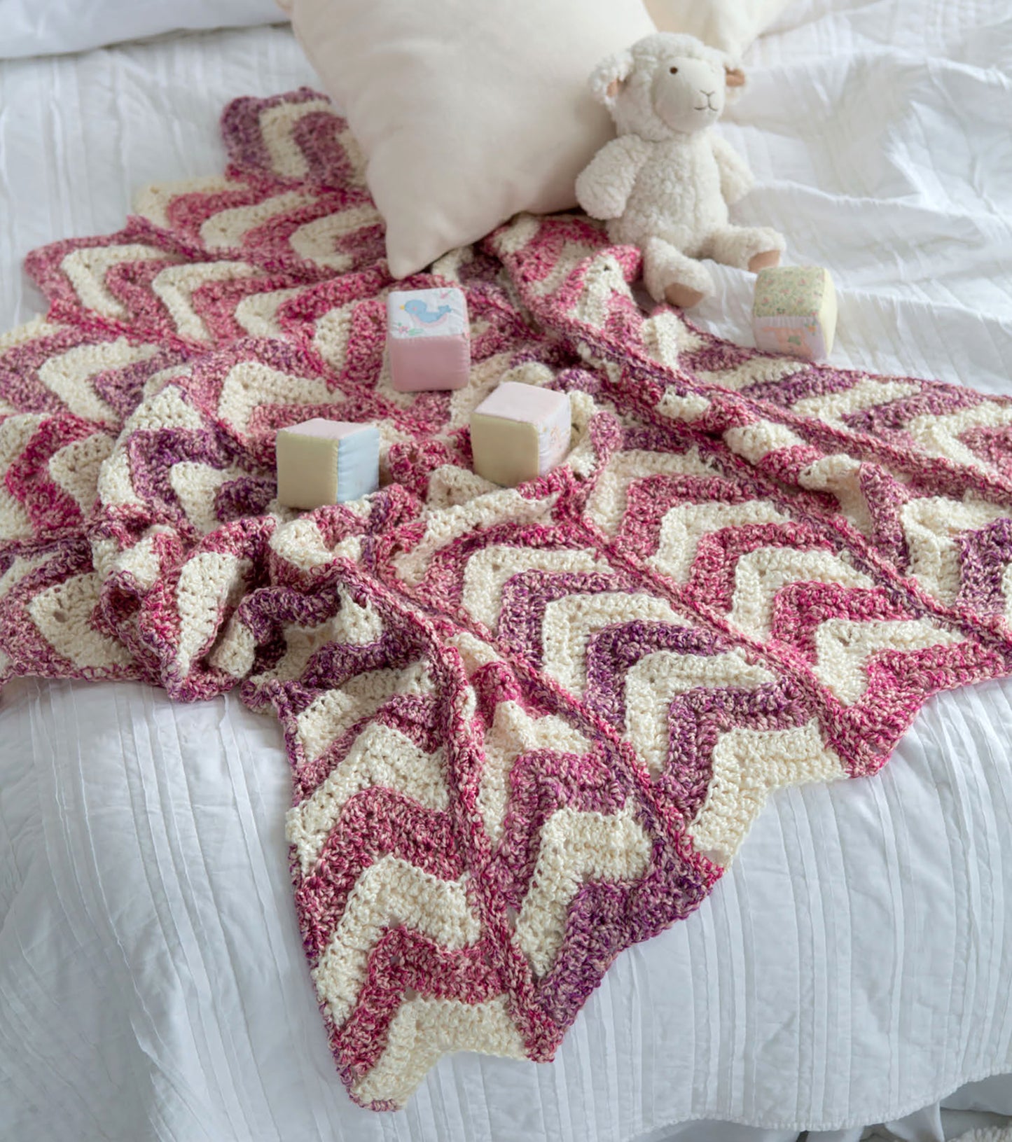 The Homespun Comfort Afghans by Leisure Arts, in pink, purple, and cream waves, lays on a white bed topped with a stuffed lamb, white pillow, and pastel toy blocks—ideal for cozy crochet throw patterns.