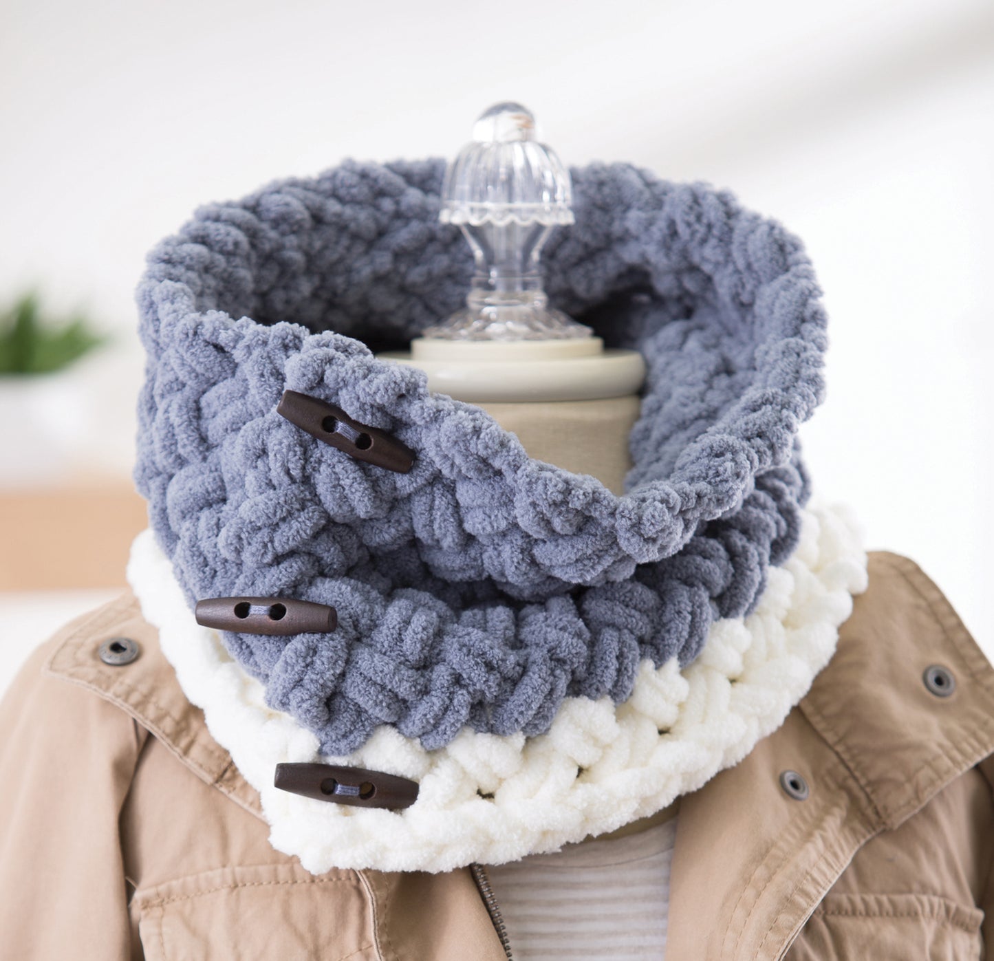 A mannequin showcases a blue and white chunky knit cowl scarf with dark brown toggles, styled with a tan jacket—an ideal beginner project featured in Leisure Arts' "Loops & Twists—Knitting Without Needles.