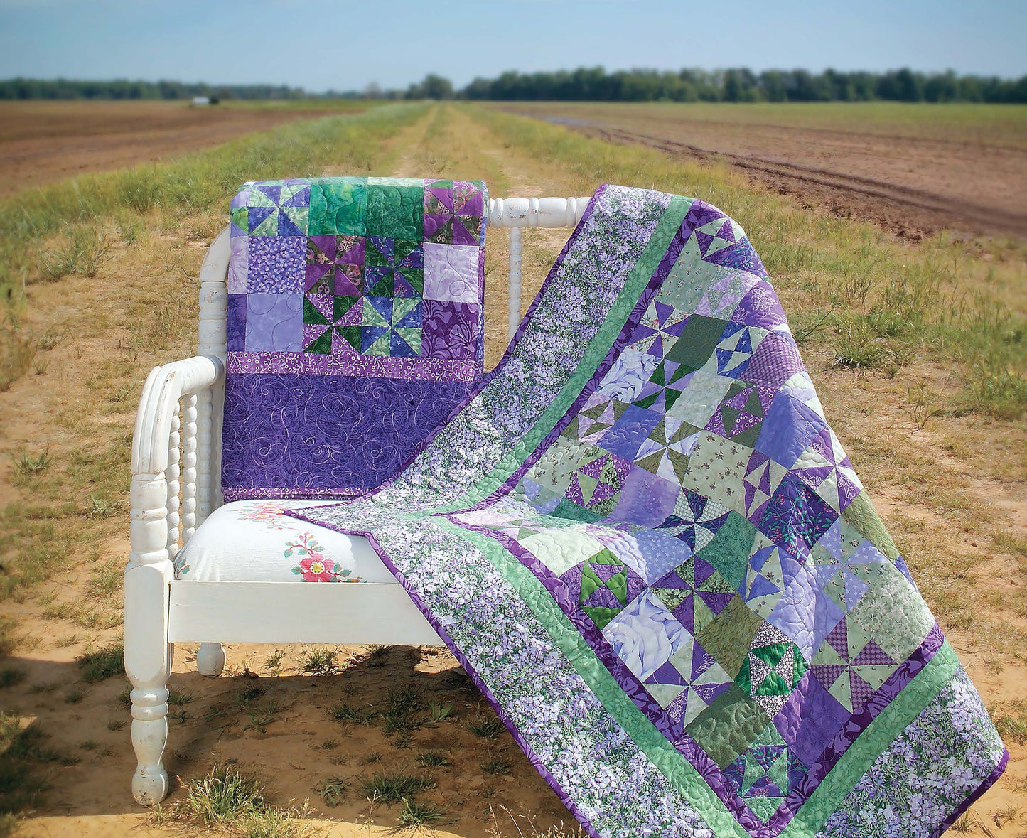 A white wooden bench sits on a dirt road, draped with vibrant triangle-patterned quilts in purple, green, and blue. The scene highlights Leisure Arts’ "It’s Quilt Magic" digital download—perfect beginner quilting inspiration.