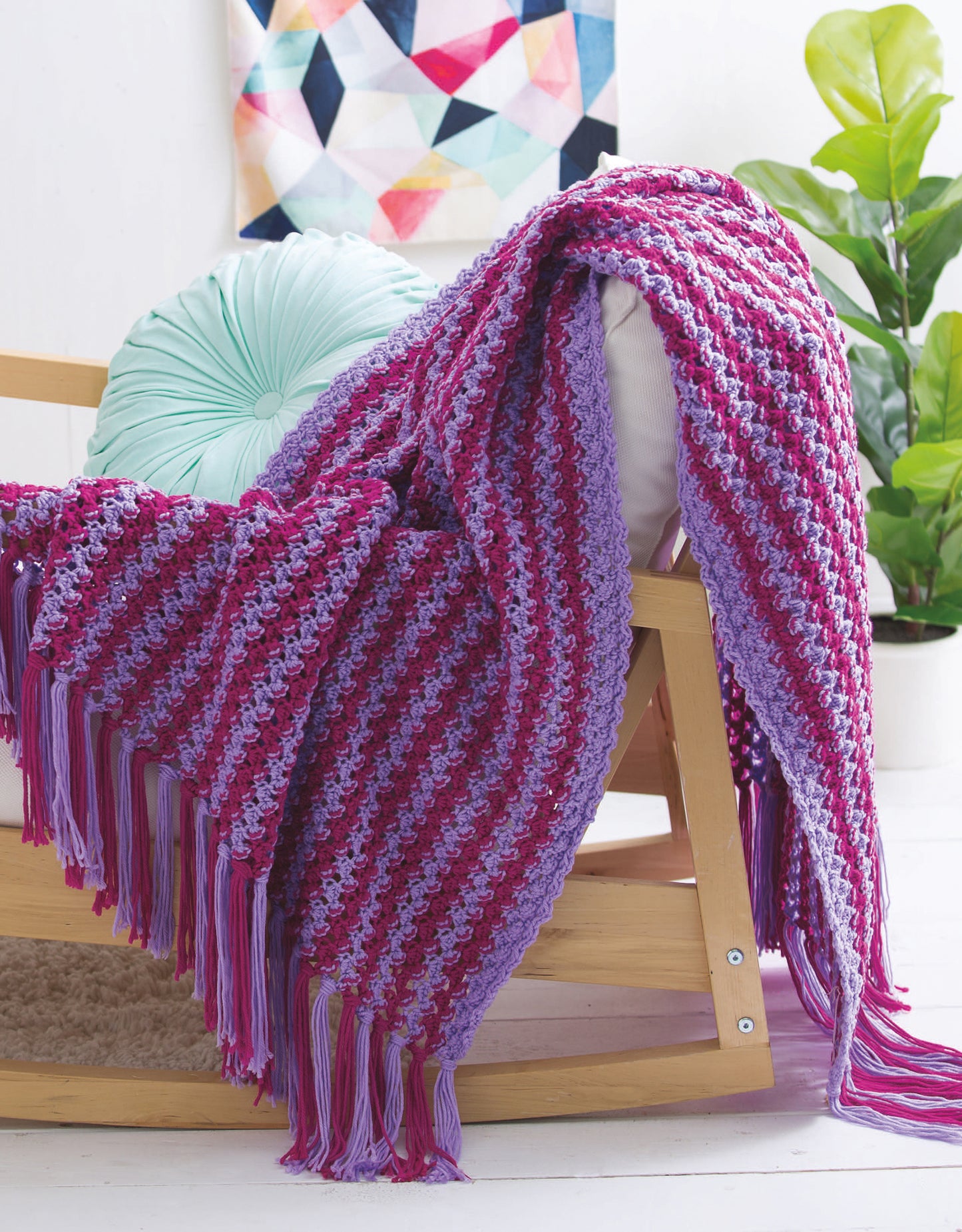 The Bright Afghans Crochet by Leisure Arts features a purple and pink striped blanket, draped over a wooden chair with a round mint-green pillow; leafy plants and abstract artwork add to the cozy atmosphere.