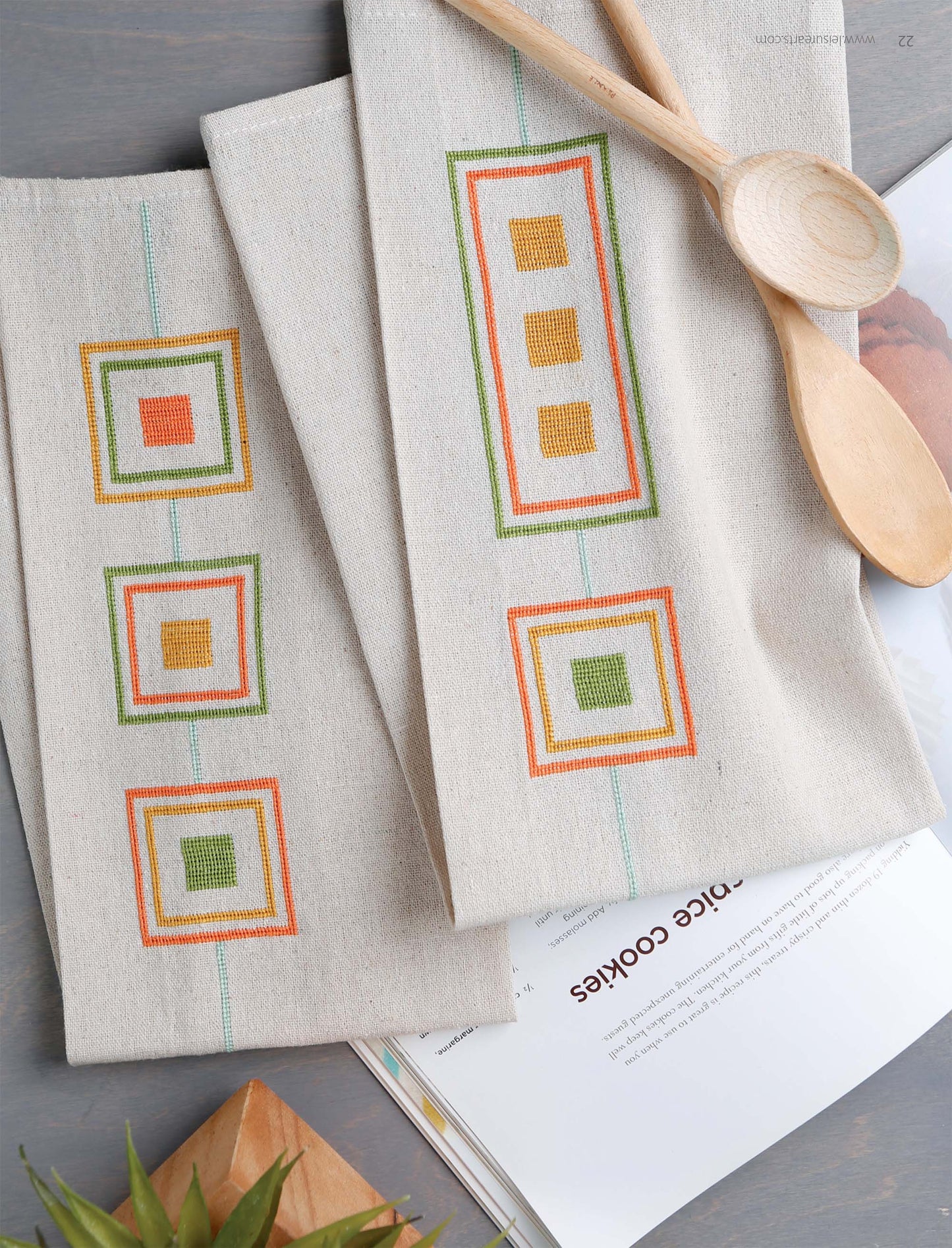 Two beige linen towels with colorful geometric embroidery, ideal for cross stitchers of all levels, lie beside spoons and a cookbook. Create your own with the "Modern Art Stitching Digital Download" by Leisure Arts.