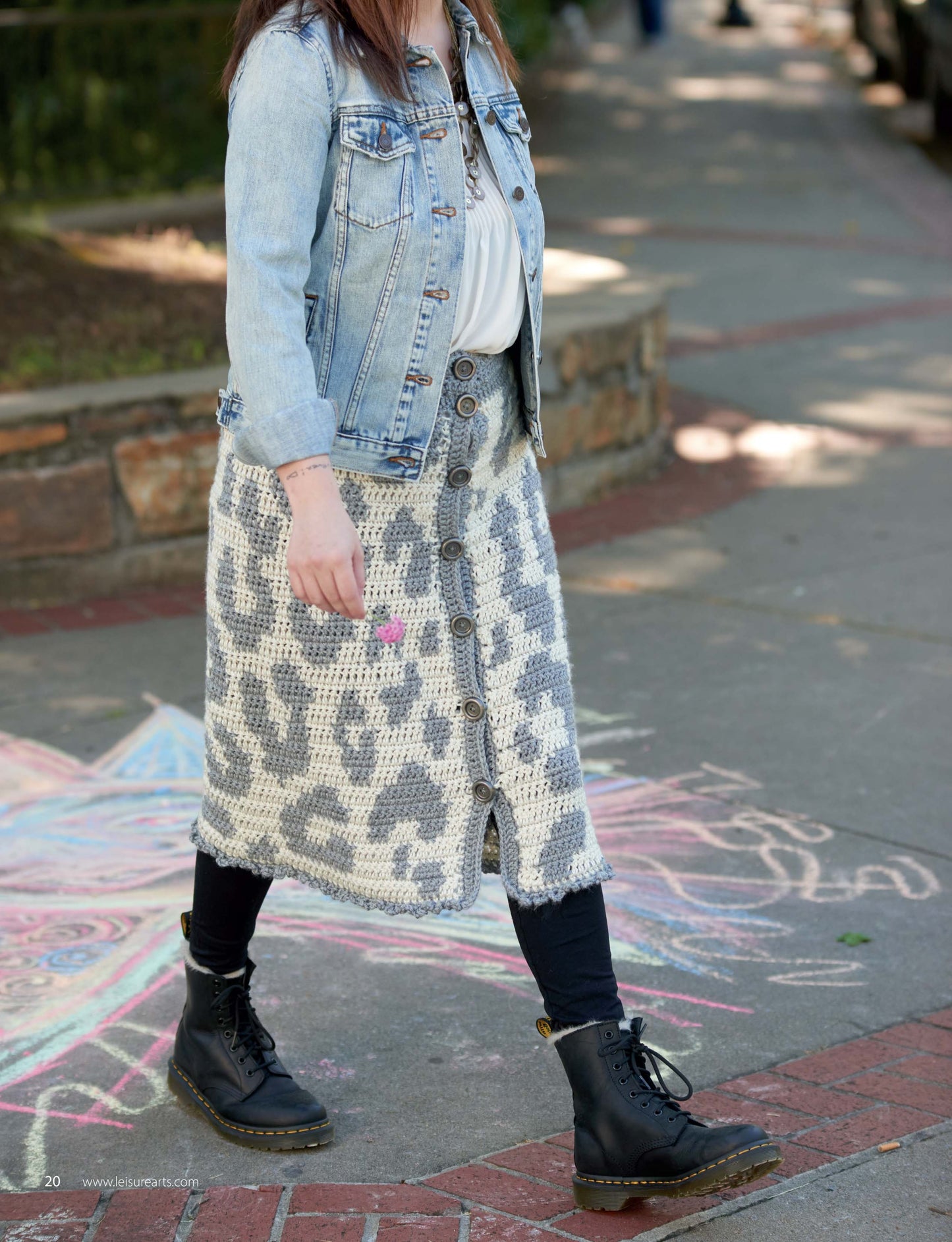 A person walks on a sidewalk with colorful chalk art, wearing a light denim jacket, white top, black boots, and a gray and white crochet skirt inspired by "Animal Prints—10 Designs" from Leisure Arts; their face is not visible.