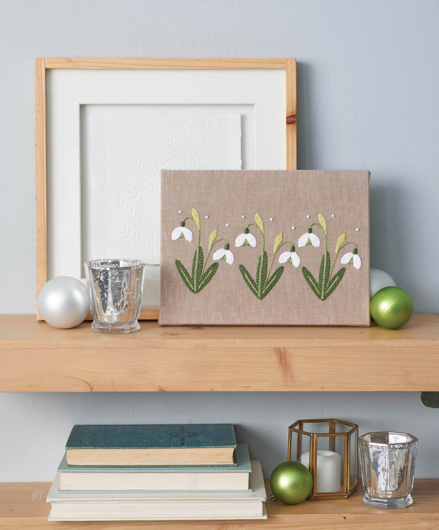 A shelf displays "Seasonal Embroidery—8 Projects to Complement What You Adore About Every Season" by Leisure Arts alongside floral linen canvas art, candle holders, ornaments, and books—perfect for craft inspiration and charming décor.