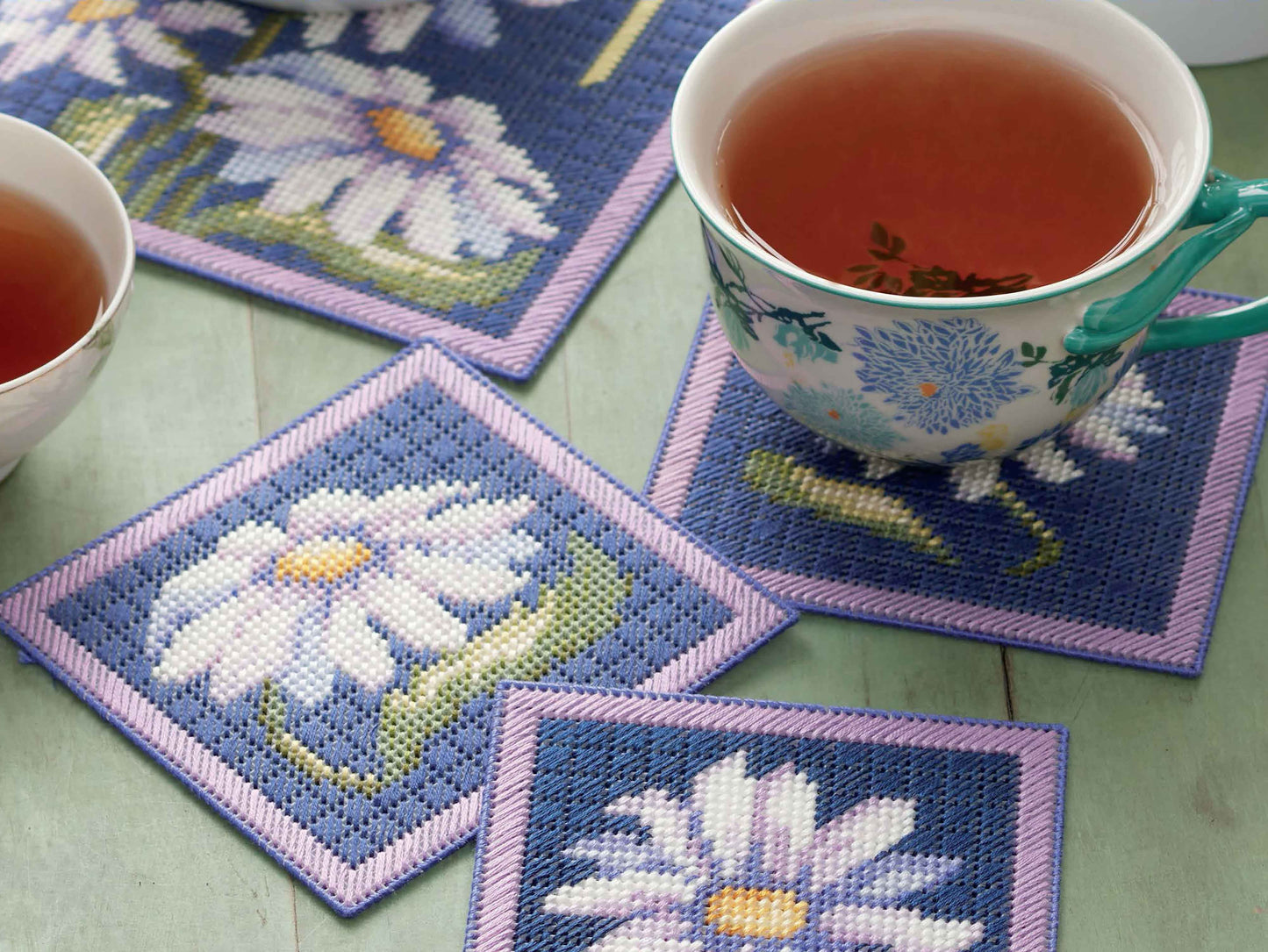 Three cups of tea sit on square "Lazy Daisies of Summer" needlepoint coasters from Leisure Arts, featuring white daisies with yellow centers and green stems on blue—perfect for beginner embroidery or plastic canvas decor projects.