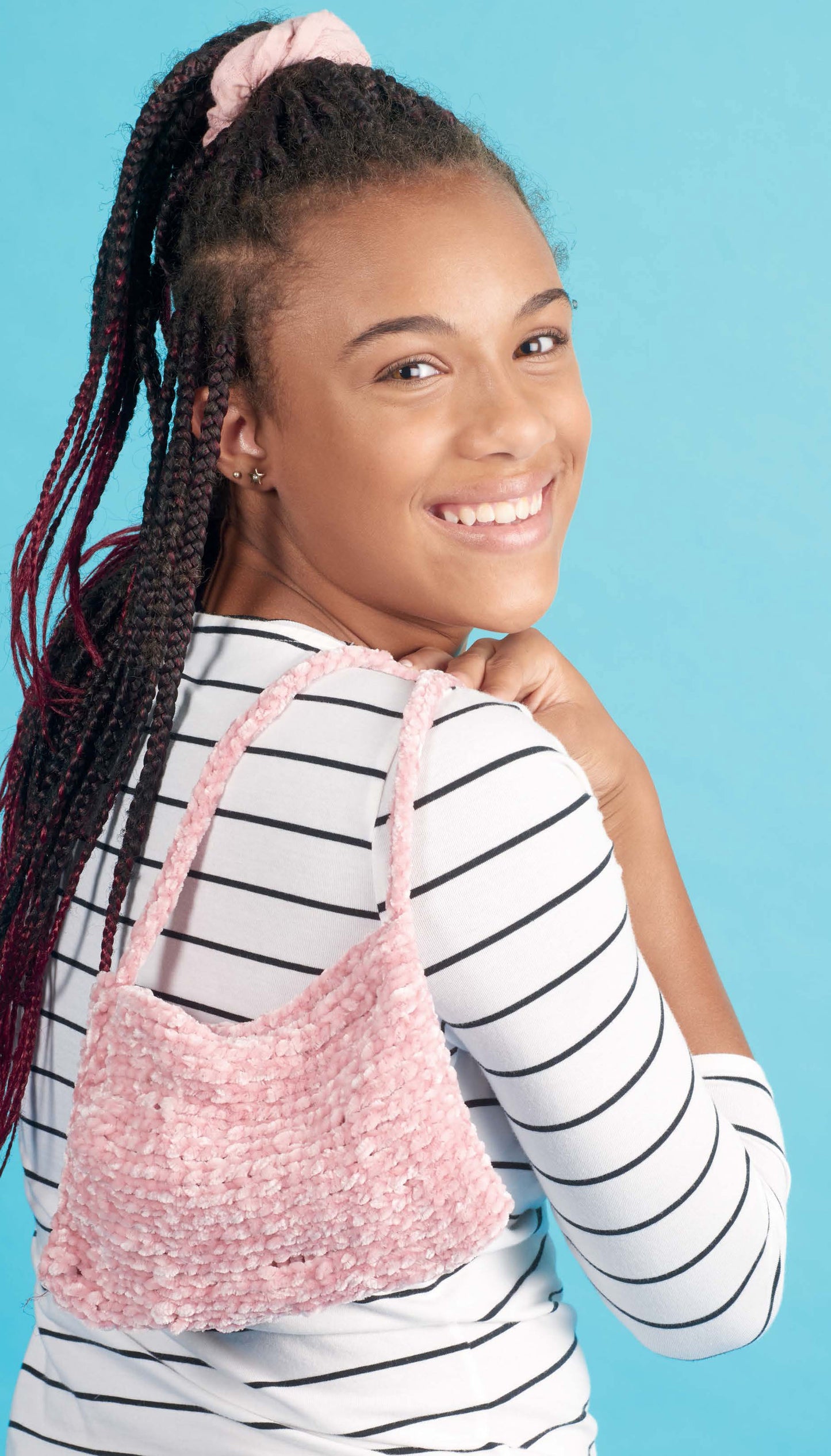 A smiling young woman with braided hair models a white and black striped top—ideal for "Beginner Knitting with Shayna Anne Rose" by Leisure Arts—carrying a pink textured handbag against a bold blue background.