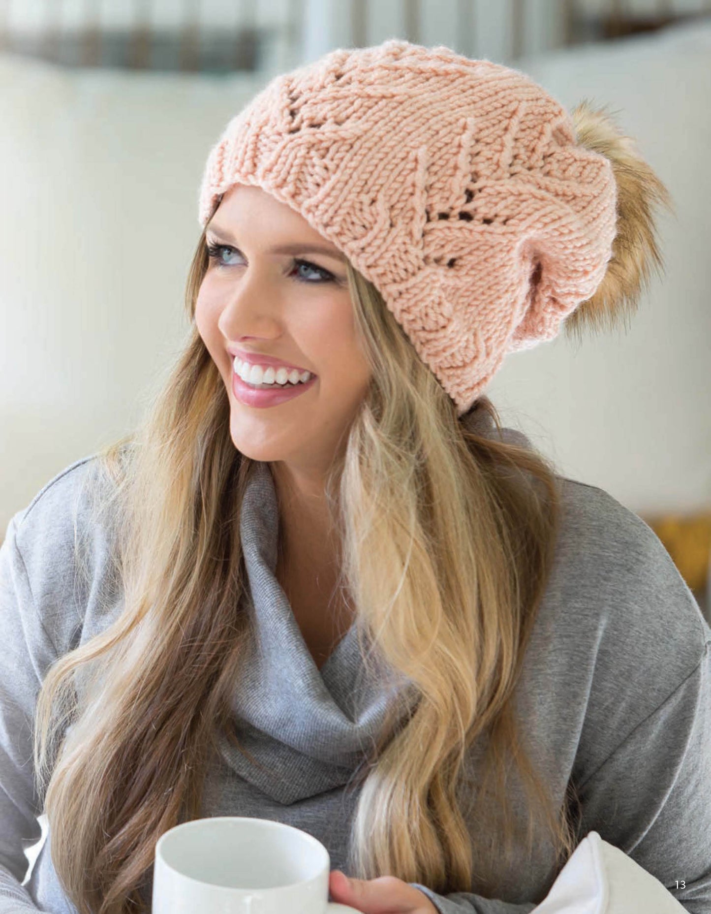 A woman with long blonde hair wears a pink knit beanie and gray sweater while holding a white mug, showcasing cozy shawls and scarves perfect for Leisure Arts' Lace Essentials in a bright indoor setting.