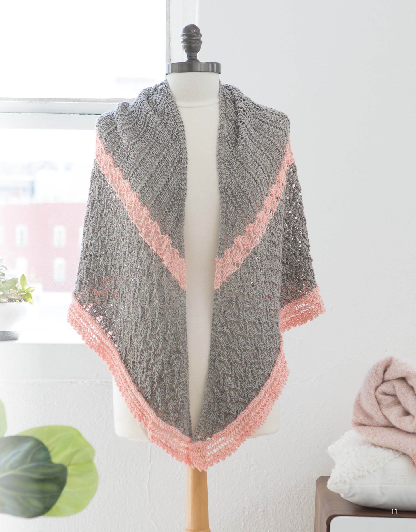 A mannequin showcases the Leisure Arts "Wrapped in Lace" shawl, gray with pink trim and delicate lace patterns, draped over its shoulders in a bright room with a window, plant, and folded blankets in the background.