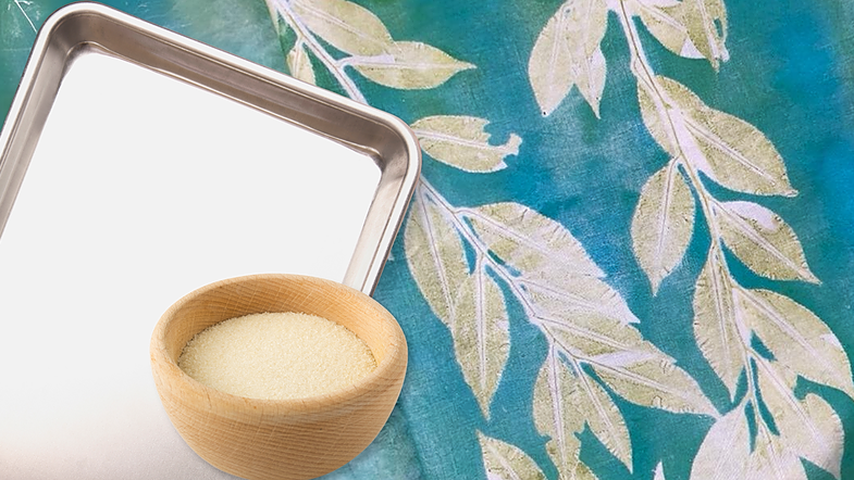 A wooden bowl of granules sits by a metal tray on vibrant fabric with white leaf patterns over teal and green. The scene evokes Creative Sparks Make Your Own Gel Printing Plate, showcasing fabric panels in harmonious colors and textures.