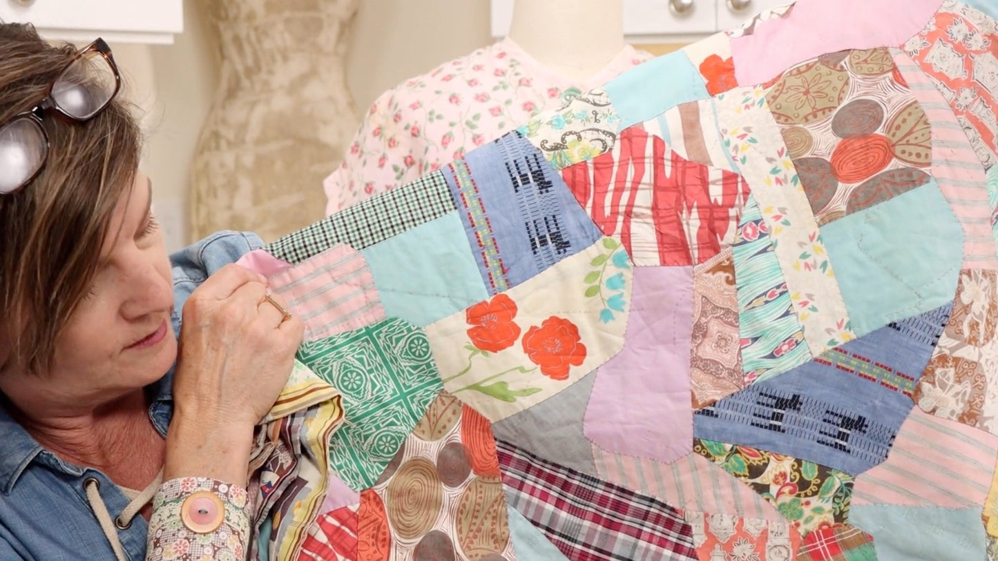 A woman admires the Feedsack Primer quilt by Creative Spark, showcasing florals, stripes, plaids, and vintage feedsack prints in a cozy room—honoring the tradition of practical projects.