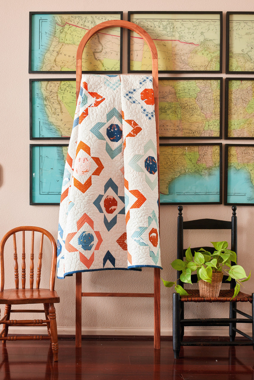 A vibrant geometric lap quilt from C&T Publishing’s Fast & Fun Lap Quilts, made with 10" squares, hangs on a wooden ladder. Nearby are two chairs and a potted plant; black-framed vintage maps decorate the beige wall behind.