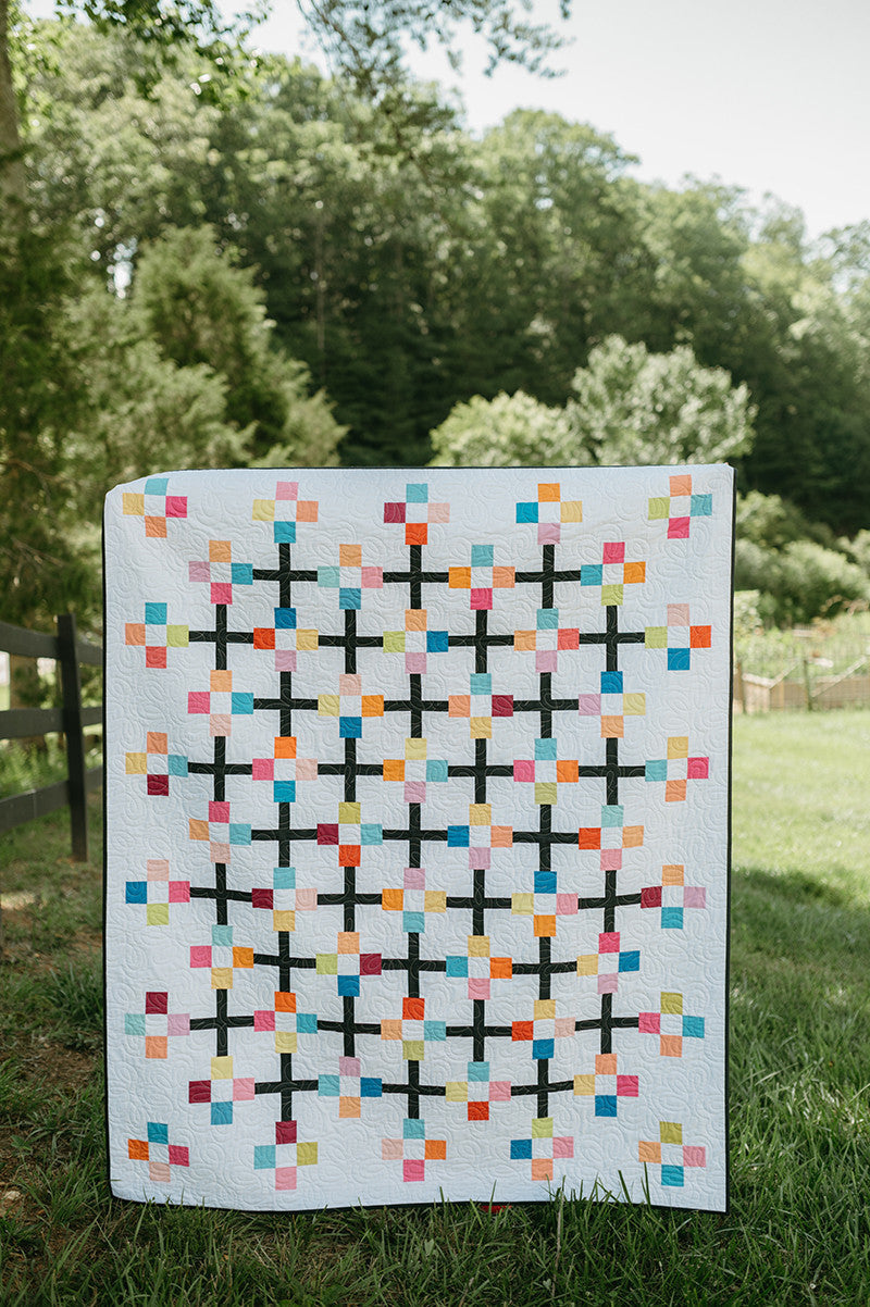 Displayed outdoors on a grassy lawn with trees and a fence, "Just Two Charm Pack Quilts" by Stash Books features a vibrant patchwork of multicolored squares and black crosses inspired by geometric quilt designs.