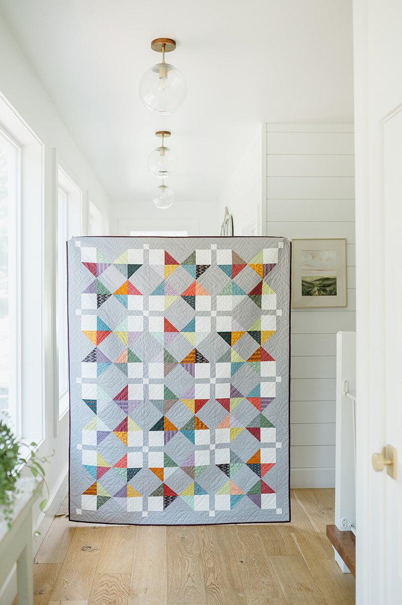 A vibrant geometric quilt from "Just Two Charm Pack Quilts" by Stash Books hangs in a bright hallway with wooden floors, white walls, and modern glass light fixtures.