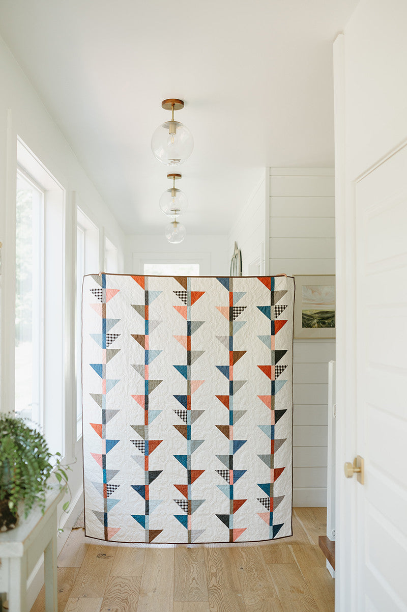 The Just Two Charm Pack Quilts by Stash Books, featuring geometric blue, orange, and black designs, hangs in a bright hallway with wood floors and white walls. Three glass pendant lights shine overhead. A green potted plant decorates a small side table nearby.