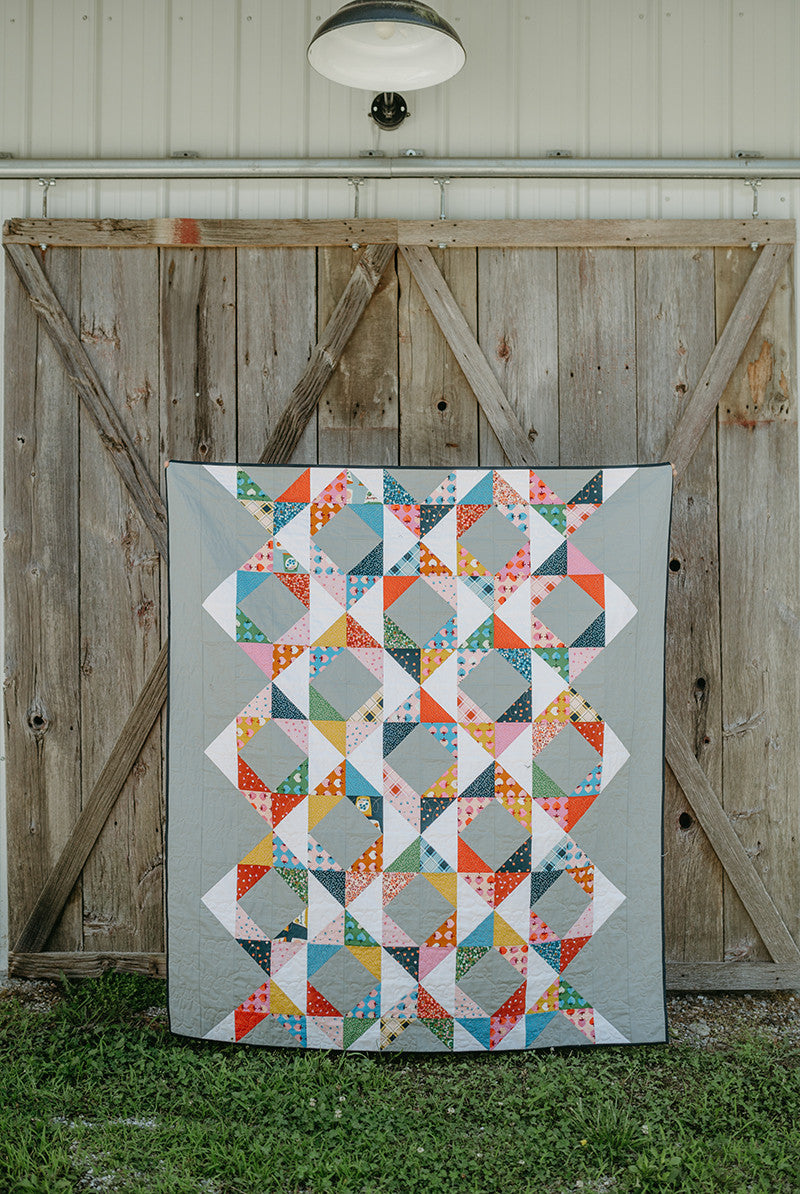 The Just Two Charm Pack Quilts by Stash Books displays vibrant geometric designs as it hangs on a rustic barn door, with grass beneath and a white outdoor lamp overhead.
