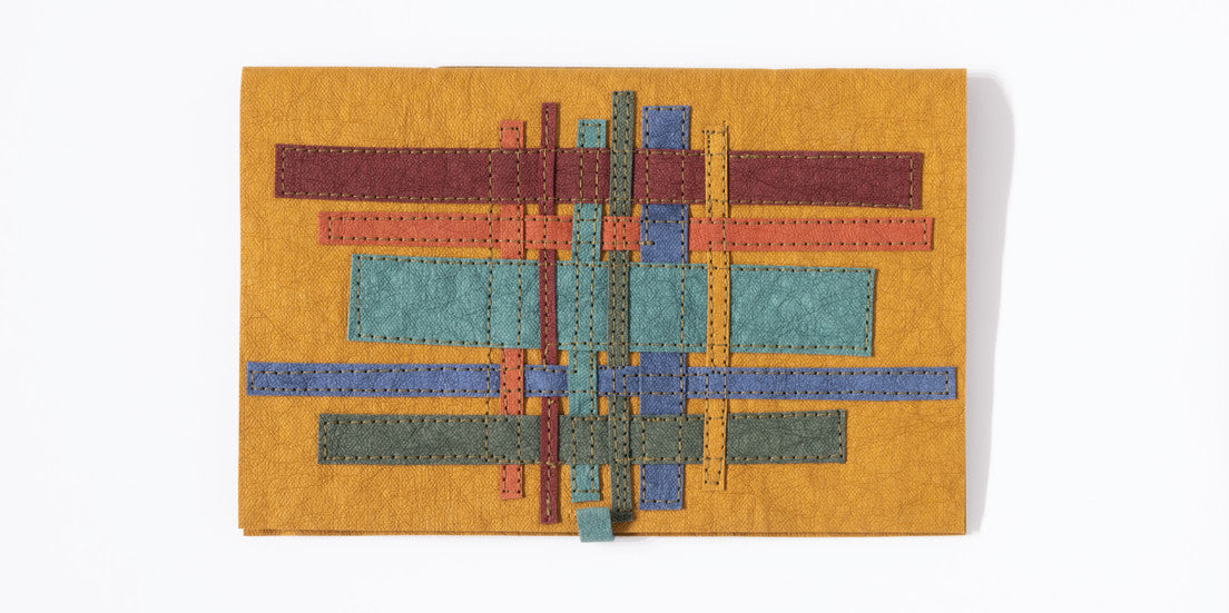 The kraft-tex Designer Sampler from C&T Publishing features a rectangular piece of mustard yellow, hand-dyed and prewashed, with stitched red, orange, blue, and green rectangles forming a bold geometric grid in this eco-friendly leather alternative.