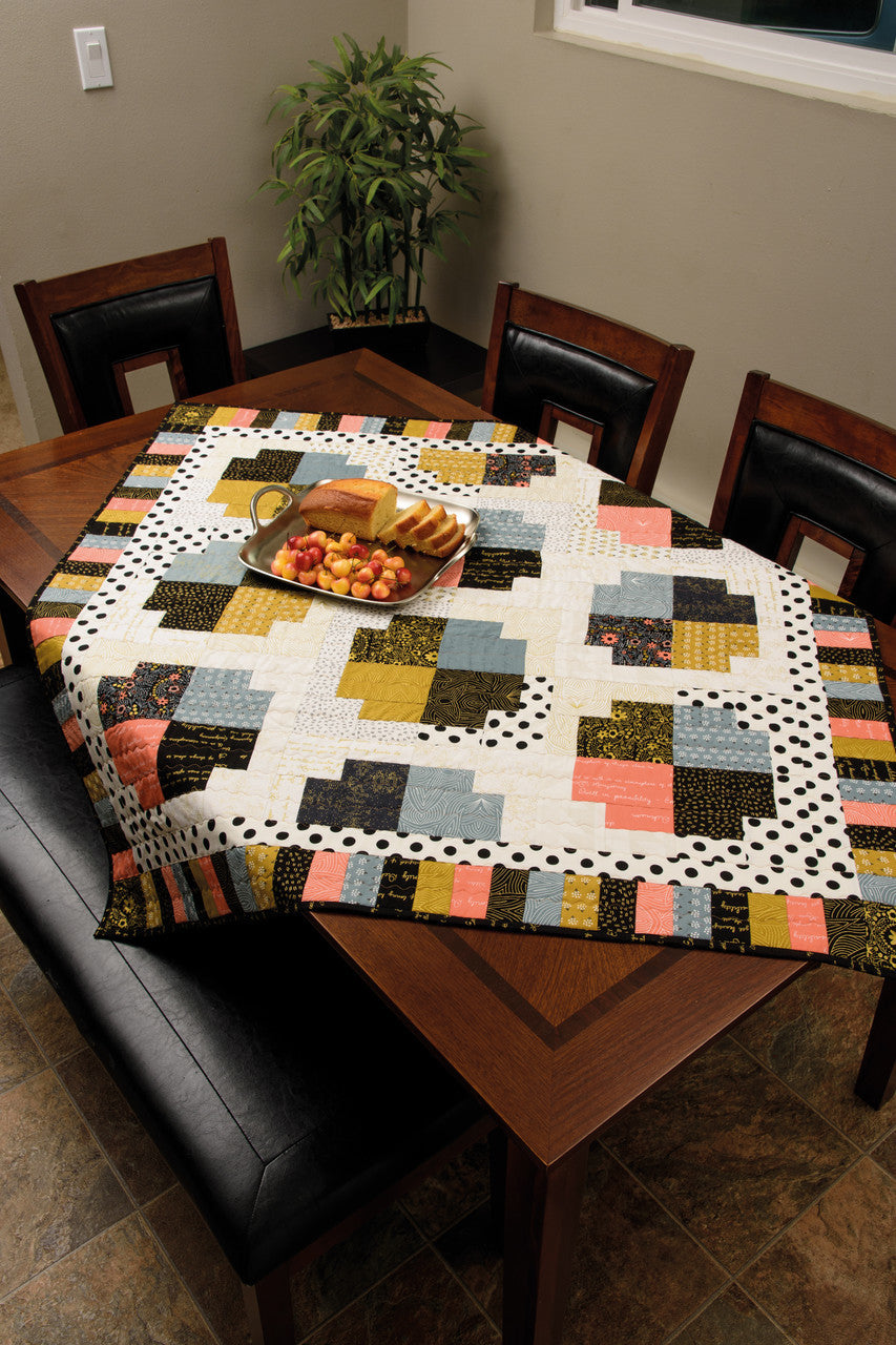 A wooden dining table features Pat Sloan's Tantalizing Table Toppers by C&T Publishing as a unique quilted topper, surrounded by four chairs. A tray with sliced bread and fruit rests on the quilt, with a potted plant near a sunlit window in the background.