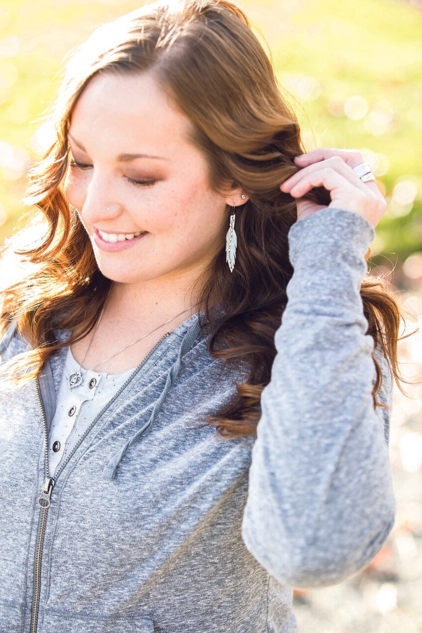 A young woman with long brown hair smiles and looks down while wearing a gray zip-up hoodie, white shirt, and DIY beaded feather earrings made using C&T Publishing’s Free Project Download: Beaded Feather Earrings. The sunlit background is softly blurred.