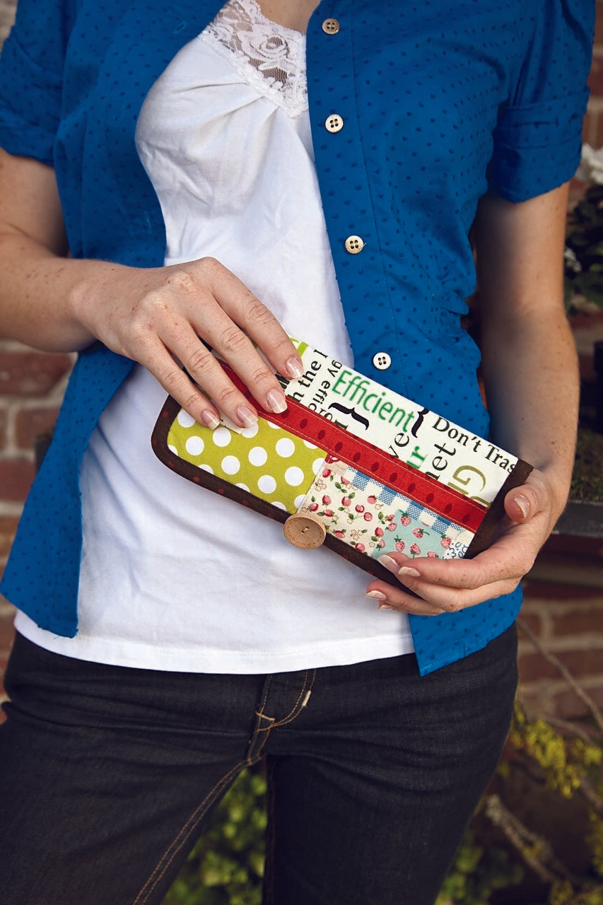 A person in a blue shirt and white top holds the colorful "Free Project Download: Betty Wallet" by C&T Publishing, featuring polka dots and text patterns, while standing in front of a brick wall.