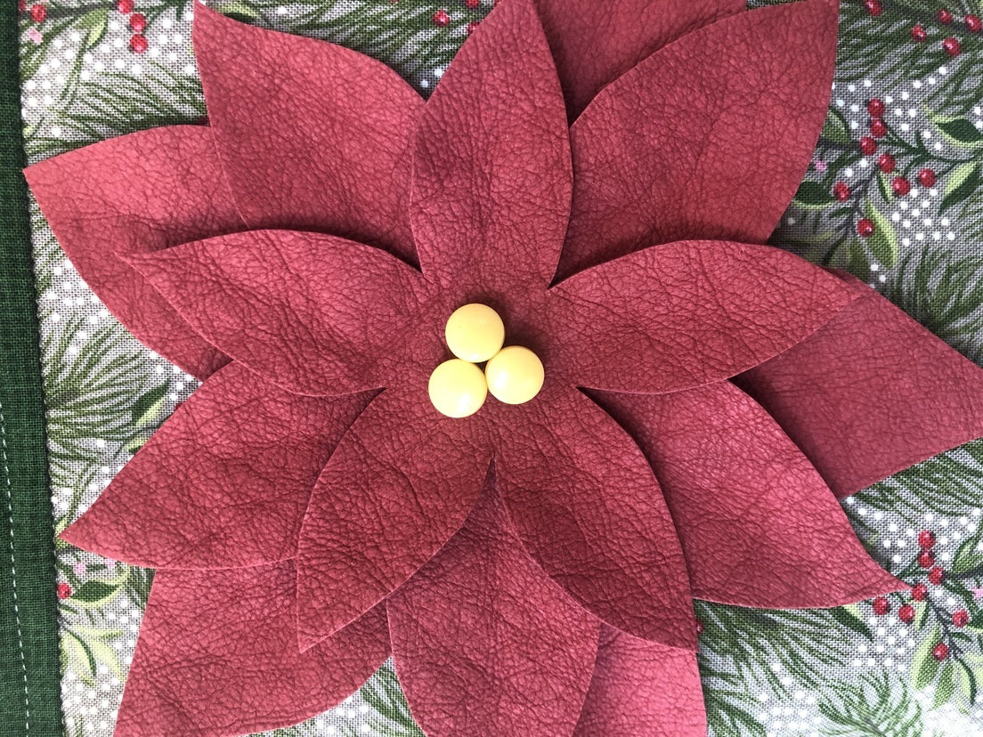A close-up of the Free Project Download: Holiday Door Stopper by C&T Publishing features layered red petals and yellow beads, set on a festive green and white background with holly, pine, and poinsettia patterns.