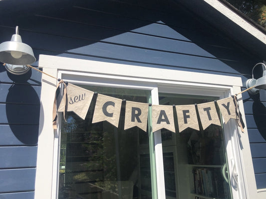 The "Free Project Download: Sew Crafty Banner" from C&T Publishing features black letters on tan kraft-tex pennants, displayed above a glass door on a blue wooden wall—ideal décor for your next sewing project.