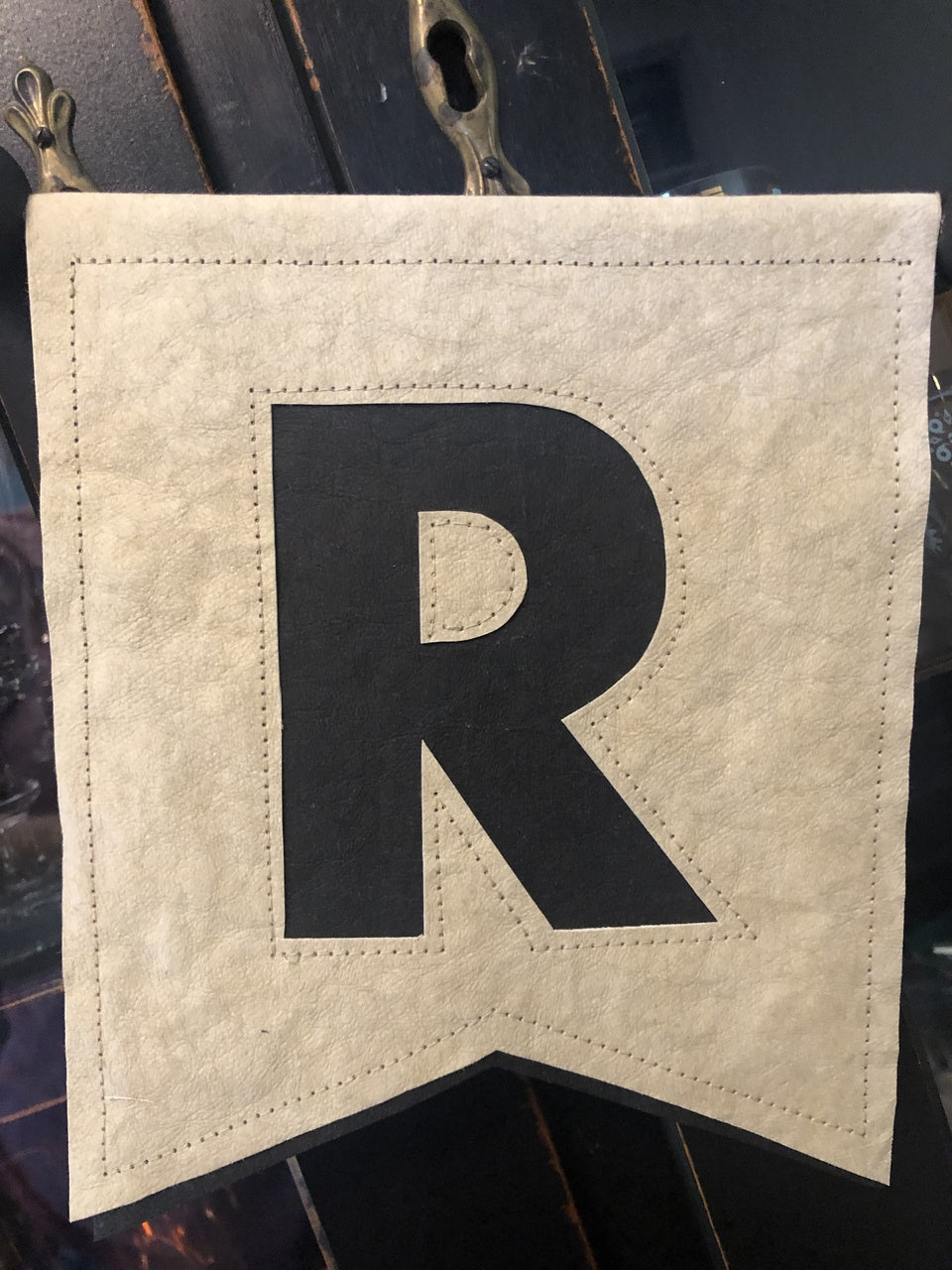 The Free Project Download: Sew Crafty Banner by C&T Publishing is a beige kraft-tex banner with a pointed bottom and bold black "R" in the center, shown hanging on dark wood—perfect for your next sewing project.