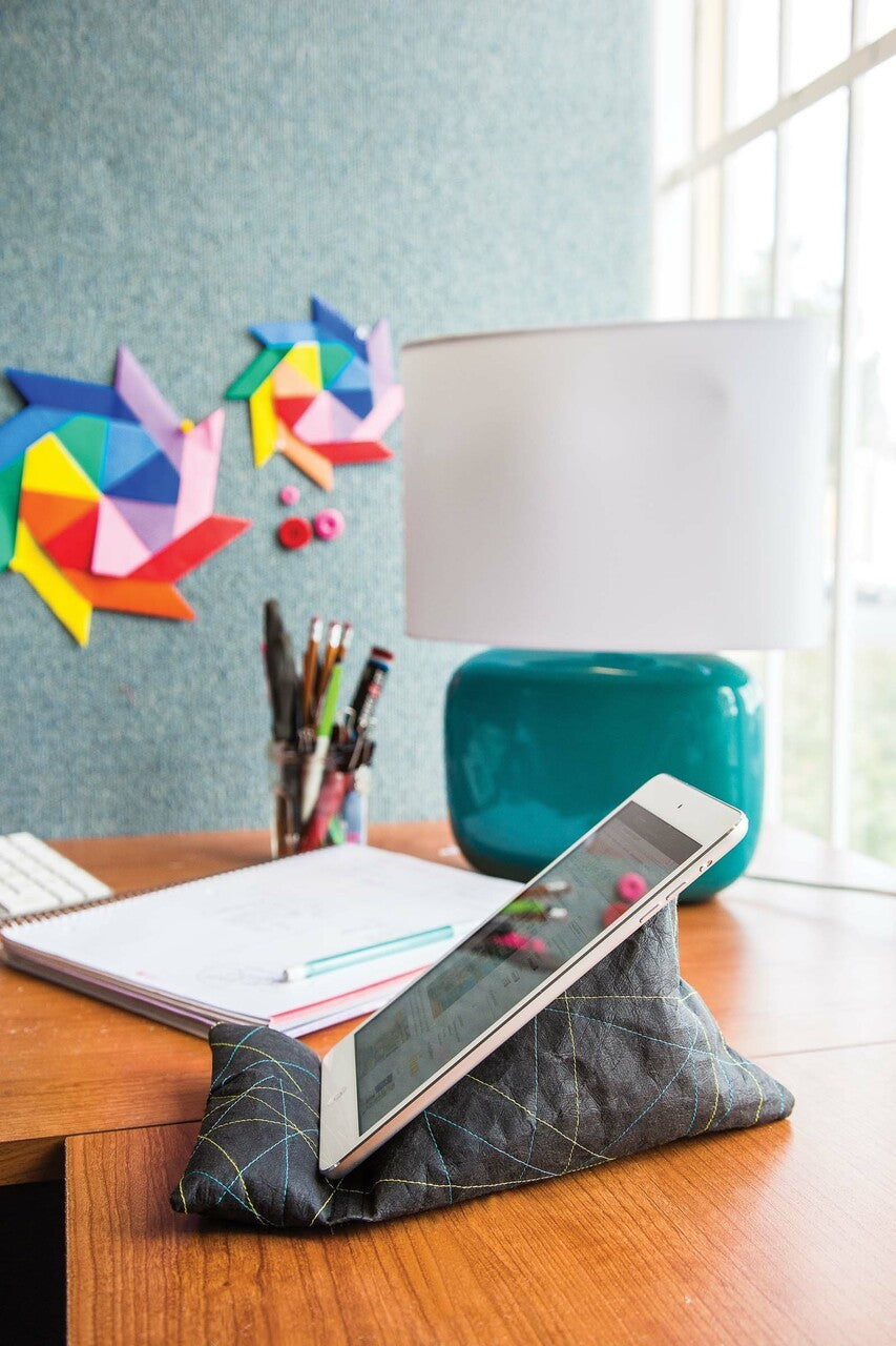 A tablet sits on the Free Project Download: Tech Stand by C&T Publishing atop a wooden desk with a lamp, notebooks, and pens. Colorful origami art decorates the wall next to a large window in the background.