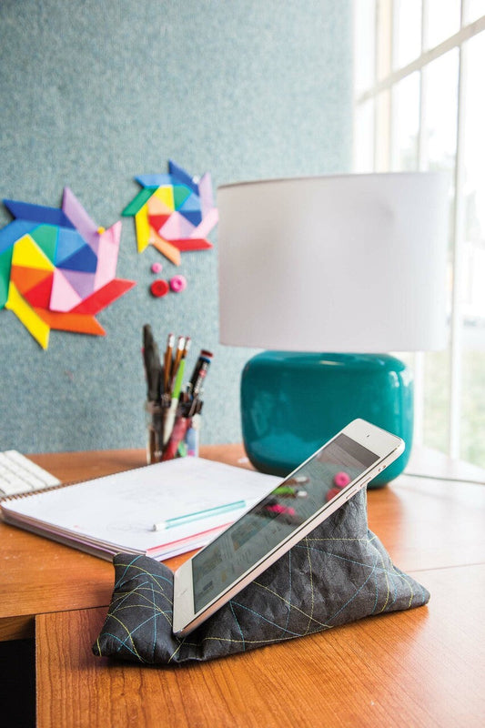 A tablet sits on the Free Project Download: Tech Stand by C&T Publishing atop a wooden desk with a lamp, notebooks, and pens. Colorful origami art decorates the wall next to a large window in the background.