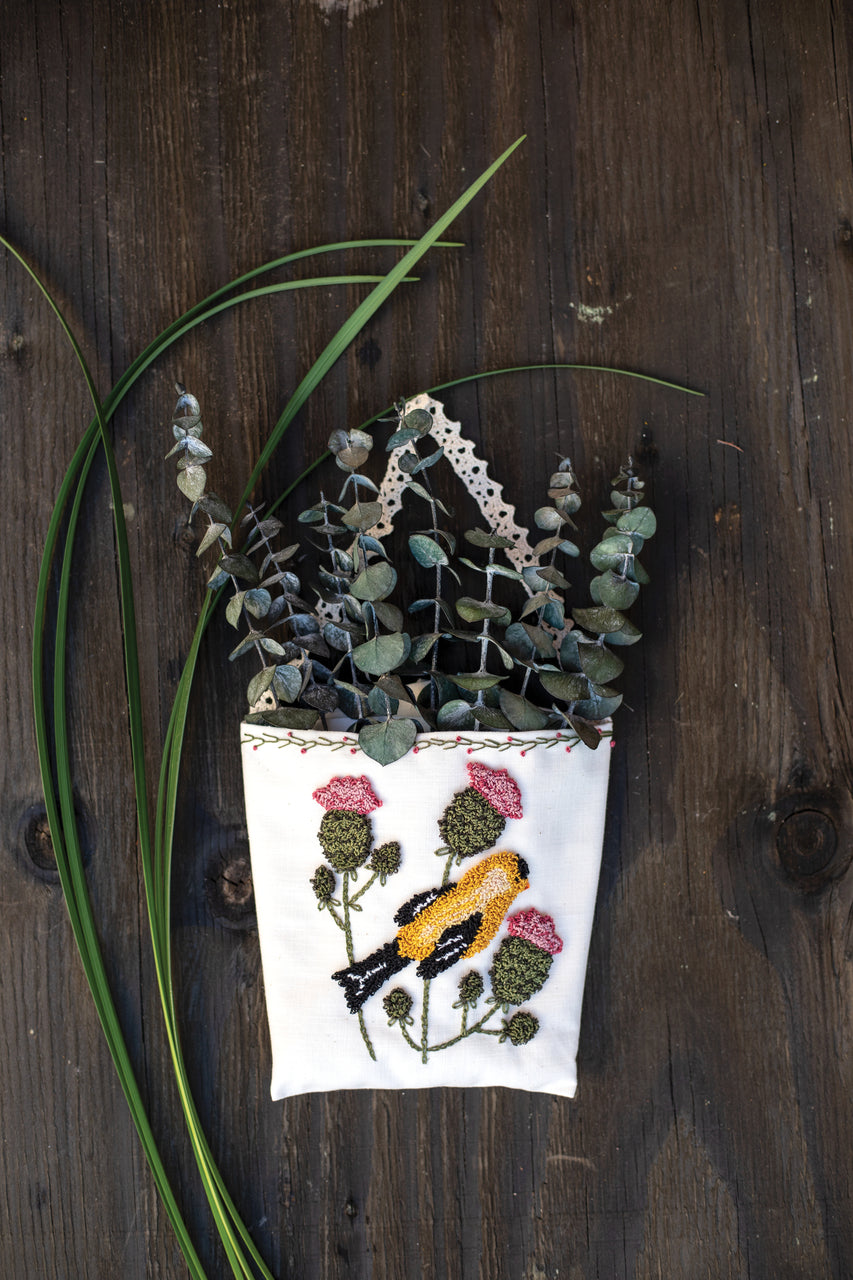 The "Jump Into Punch Needle" pouch by C&T Publishing, featuring embroidered pink flowers and a yellow bird, is filled with eucalyptus branches and hangs on a rustic wood wall with long green leaves draping beside it.