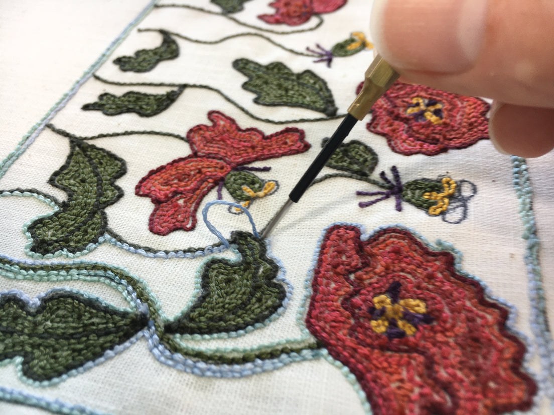 A close-up of a hand using Jump Into Punch Needle by C&T Publishing to create vibrant red flowers, green leaves, and bees on white fabric—an ideal fiber art project for beginners.