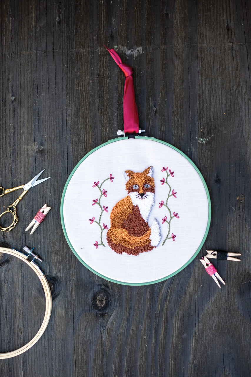 Jump Into Punch Needle by C&T Publishing features a stitched fox with pink flowers on white fabric, displayed on a dark wood surface with embroidery scissors, thread-wrapped clothespins, and an empty hoop—ideal for beginners.