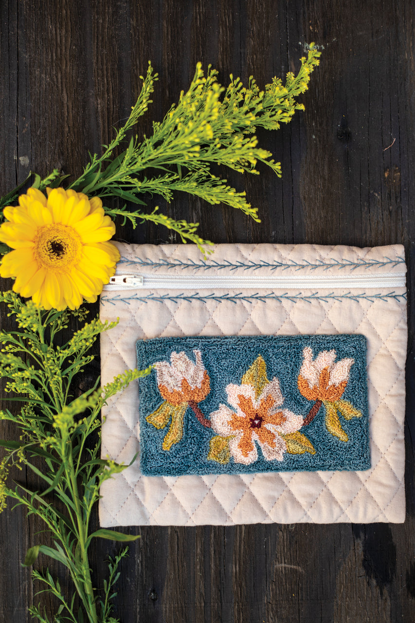The "Jump Into Punch Needle" pouch by C&T Publishing features cream quilted fabric with blue stitching and floral punch needle embroidery, displayed on dark wood and accented by yellow flowers—an ideal fiber art project for beginners.
