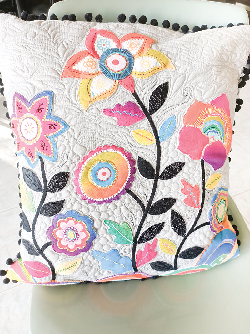 The "Machine Magic" pillow by C&T Publishing features vibrant embroidered flowers and leaves, intricate stitching on a white background, and black pom-pom trim, shown resting on a light-colored chair.