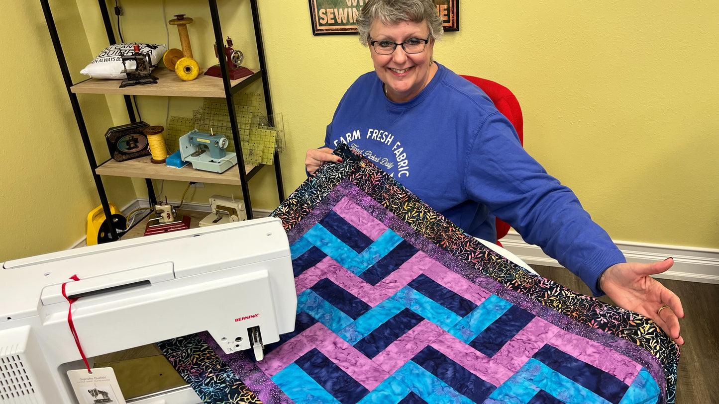 In a sewing room filled with Creative Sparks Quilting 101 essentials, a smiling person wearing glasses showcases a vibrant quilt. A sewing machine softly hums on the table, while shelves stocked with stitching supplies hint at years of expert guidance.