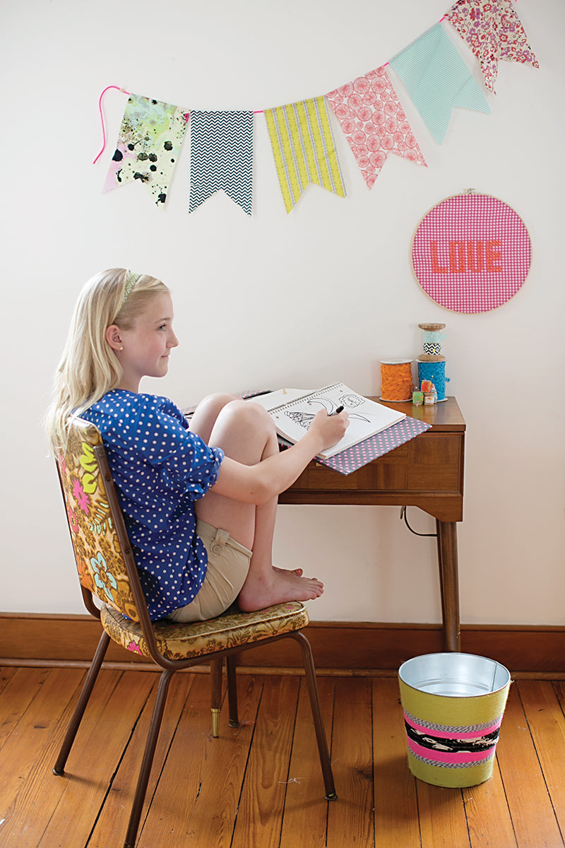 A young girl sits barefoot at a small wooden desk, drawing in a notebook—dreaming up her next My Sewing Workshop project from FunStitch Studio. Colorful fabric bunting and a pink embroidery hoop with "LOVE" decorate the wall above her.
