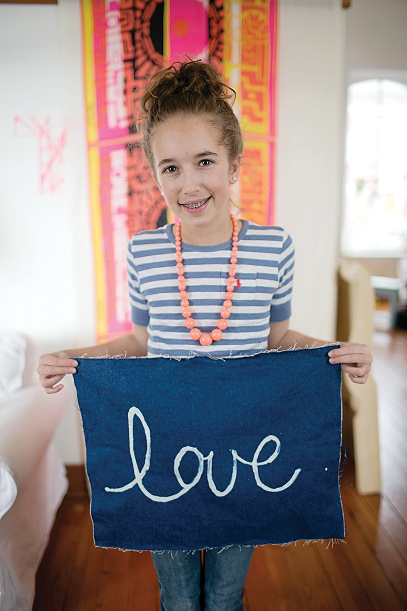 A smiling girl with curly hair holds her "love" project from My Sewing Workshop by FunStitch Studio—a fun kids crafting kit—while standing in a brightly lit, colorfully decorated room.