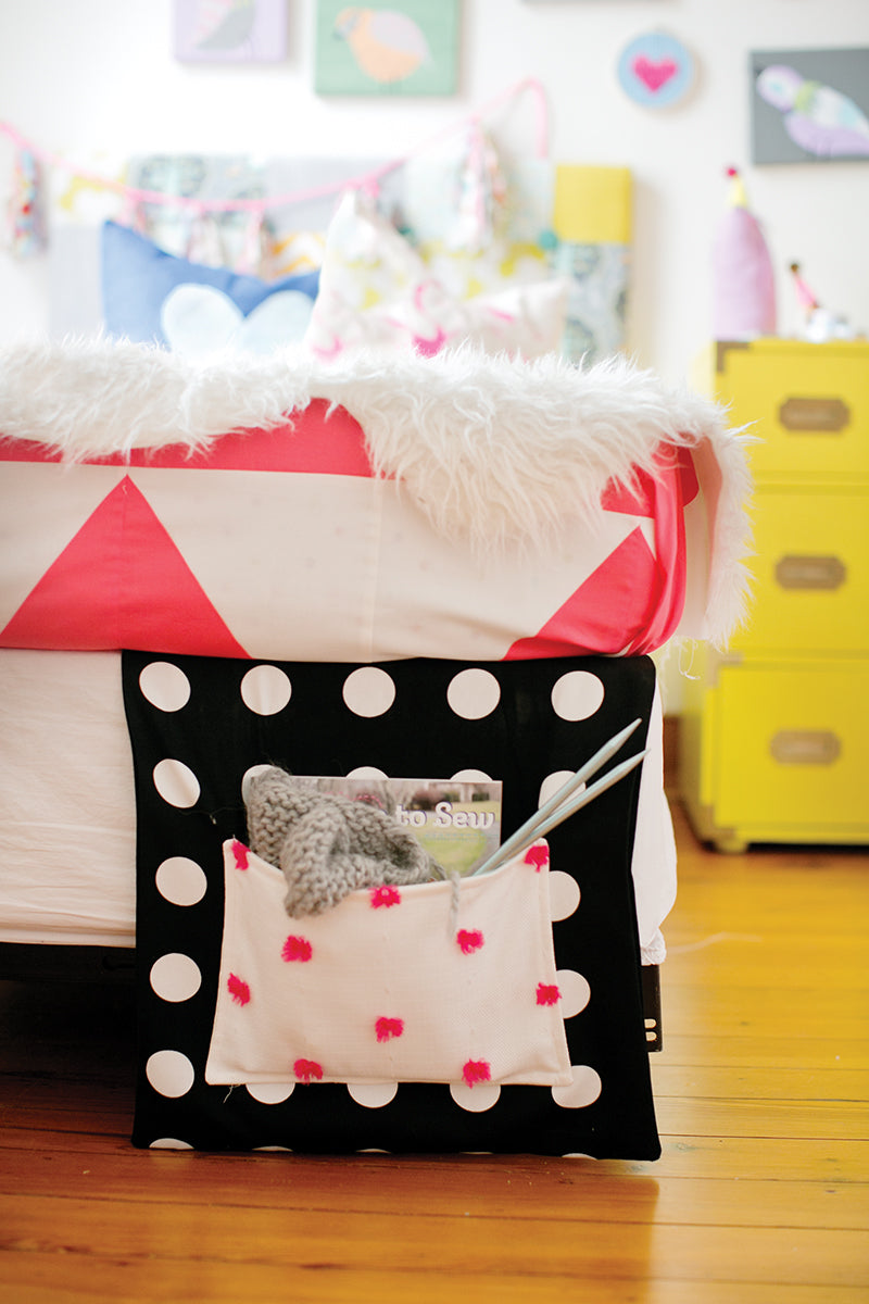 The My Sewing Workshop bedside caddy by FunStitch Studio features pink and white polka dots, perfect for kids' crafts. It hangs from a bed with a red and white blanket, holding knitting needles, yarn, and a book. A yellow dresser is in the background.