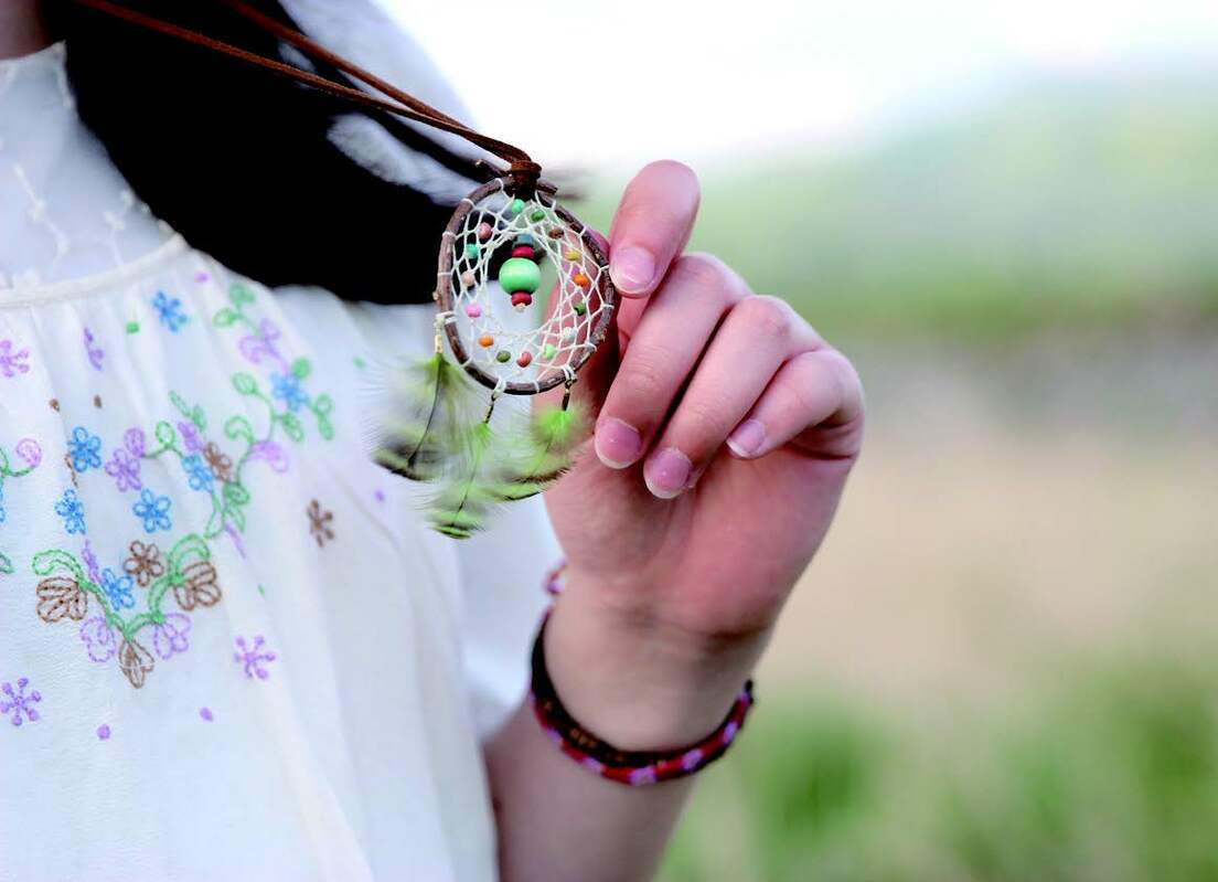 A person in a white embroidered top holds a small dreamcatcher necklace with green beads and feathers, crafted from nature-inspired materials. Featured: "Create Dream Catchers" by Stash Books. The background shows an outdoor setting.