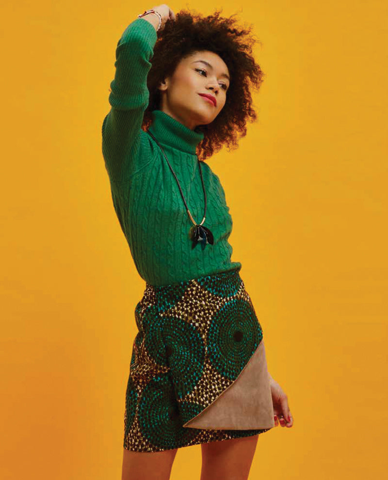 A woman with curly hair poses in a green turtleneck and an African wax print skirt, inspired by "Creating with African Wax Fabric" from Stash Books—a look celebrated by sewists and designers.