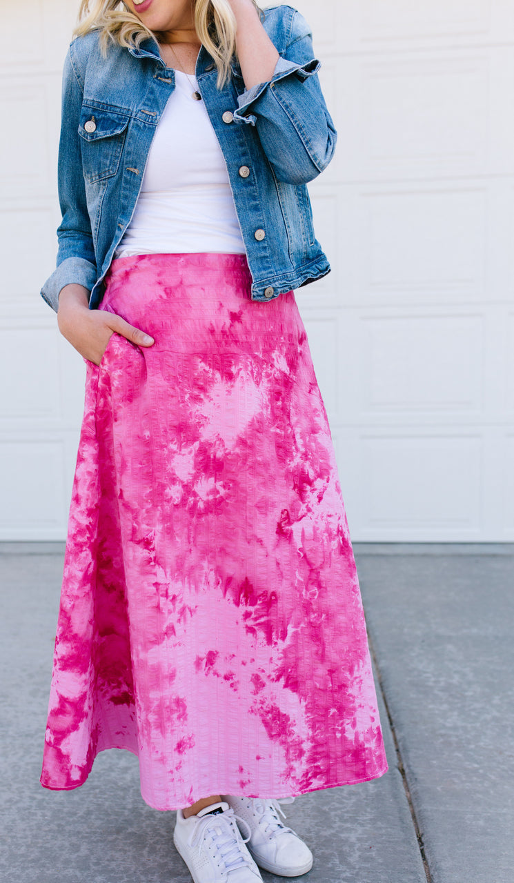 A woman in a blue denim jacket, white tee, and a pink tie-dye maxi skirt—made with step-by-step tips from "DIY Guide to Tie Dye Style" by Stash Books—stands on concrete before a white garage door, one hand in her pocket.