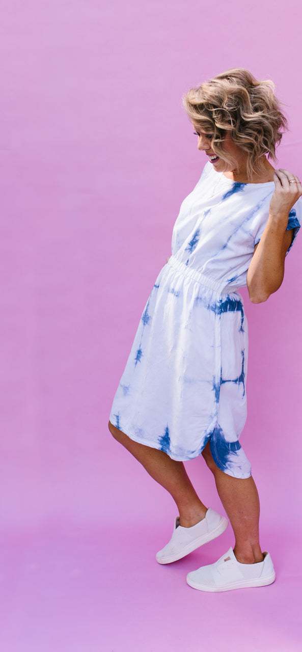 A woman with curly blonde hair wears a blue and white tie-dye dress inspired by the "DIY Guide to Tie Dye Style" from Stash Books, paired with white sneakers, standing against a pink background, looking down and holding her hair.