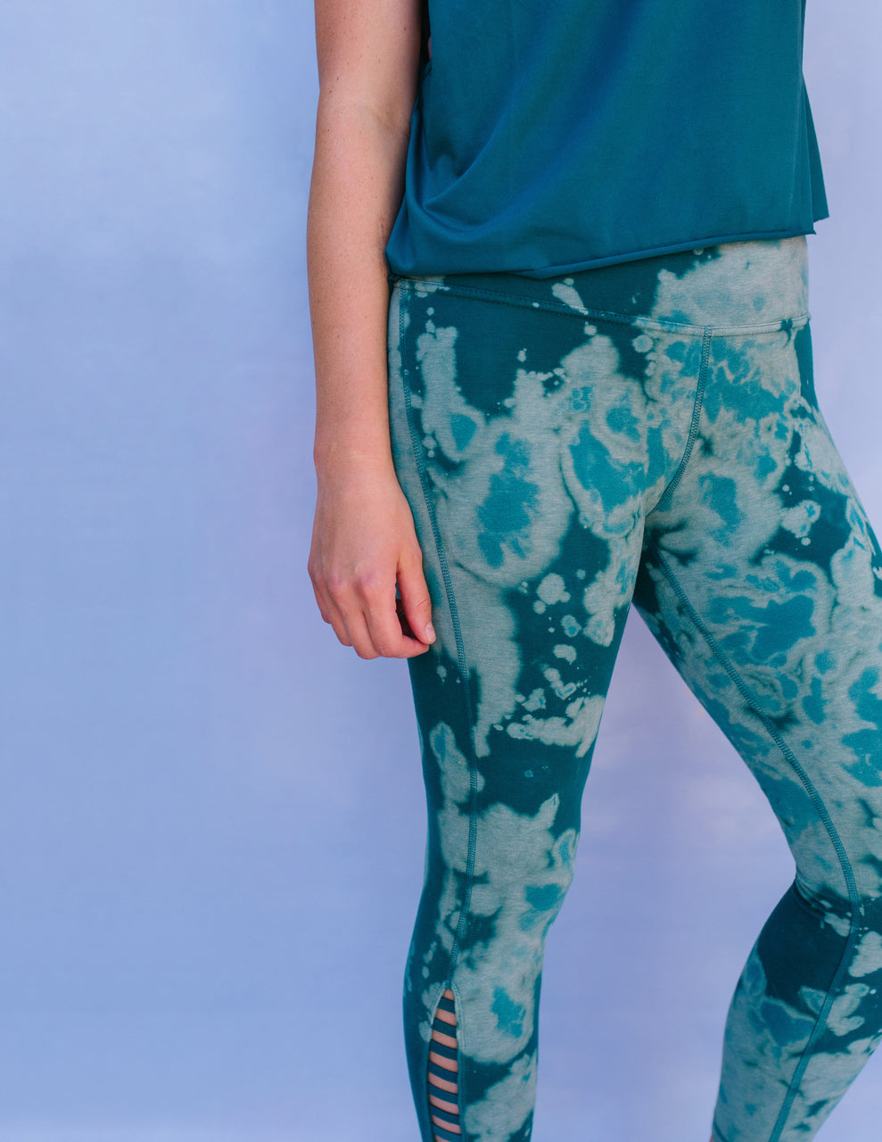 Wearing a teal top and teal tie-dye leggings created with techniques from the DIY Guide to Tie Dye Style by Stash Books, a person stands against a light blue background with only their torso, right arm, and upper legs visible.